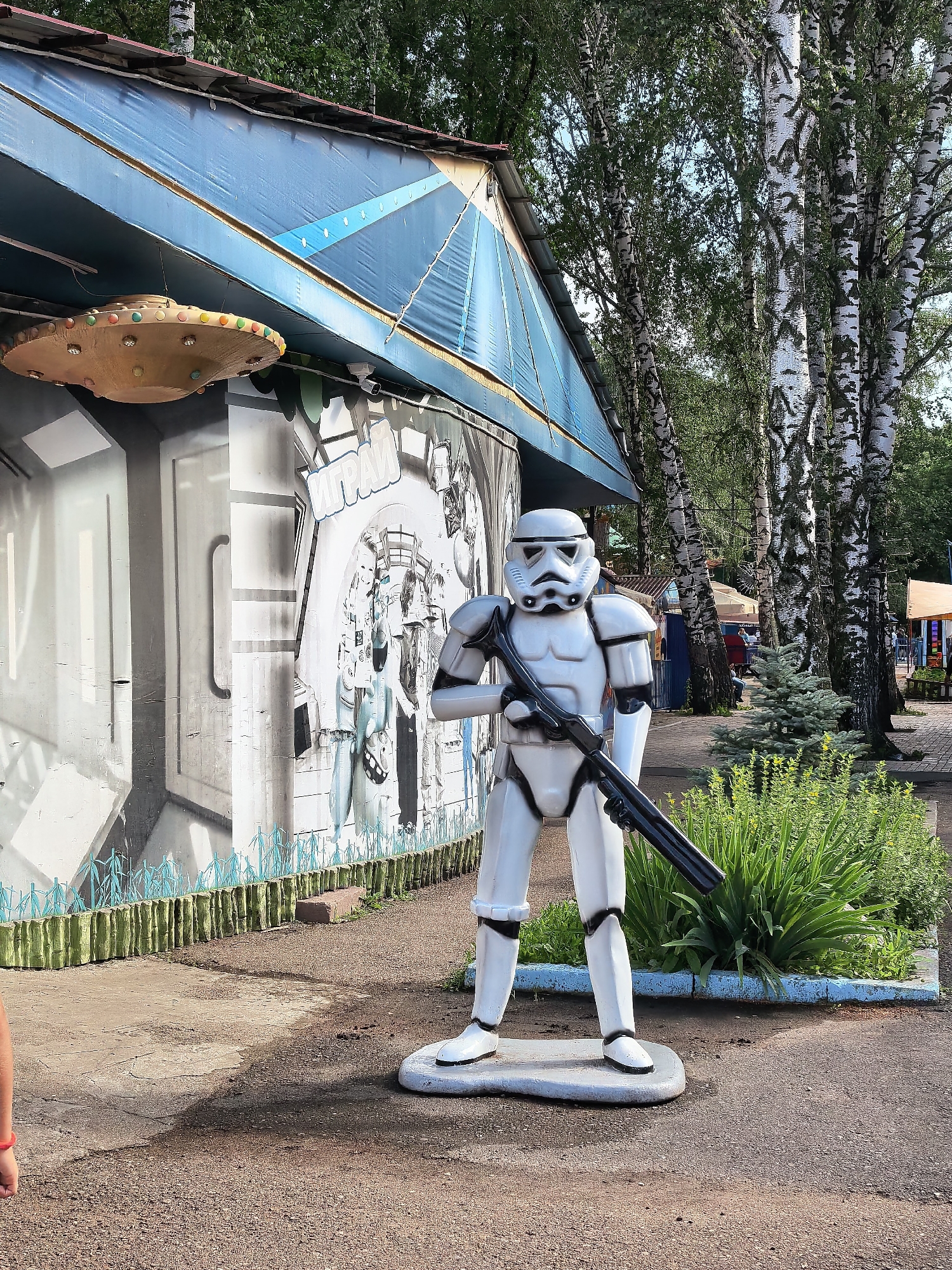 In Sterlitamak, Russia, a life-size statue of a Star Wars stormtrooper, complete with a blaster rifle, stands outdoors on a paved pathway. The statue is positioned in front of a building with a blue awning and a large mural on its exterior wall. The mural appears to be in black and white and depicts abstract shapes and figures. Hanging from the building's awning is a decorative, disc-shaped object with colorful lights. To the right of the stormtrooper statue is dense green shrubbery. In the background, several birch trees with their characteristic white bark are visible, along with other structures and trees suggesting a park or recreational area. The ground around the statue is a mix of dirt and gravel.