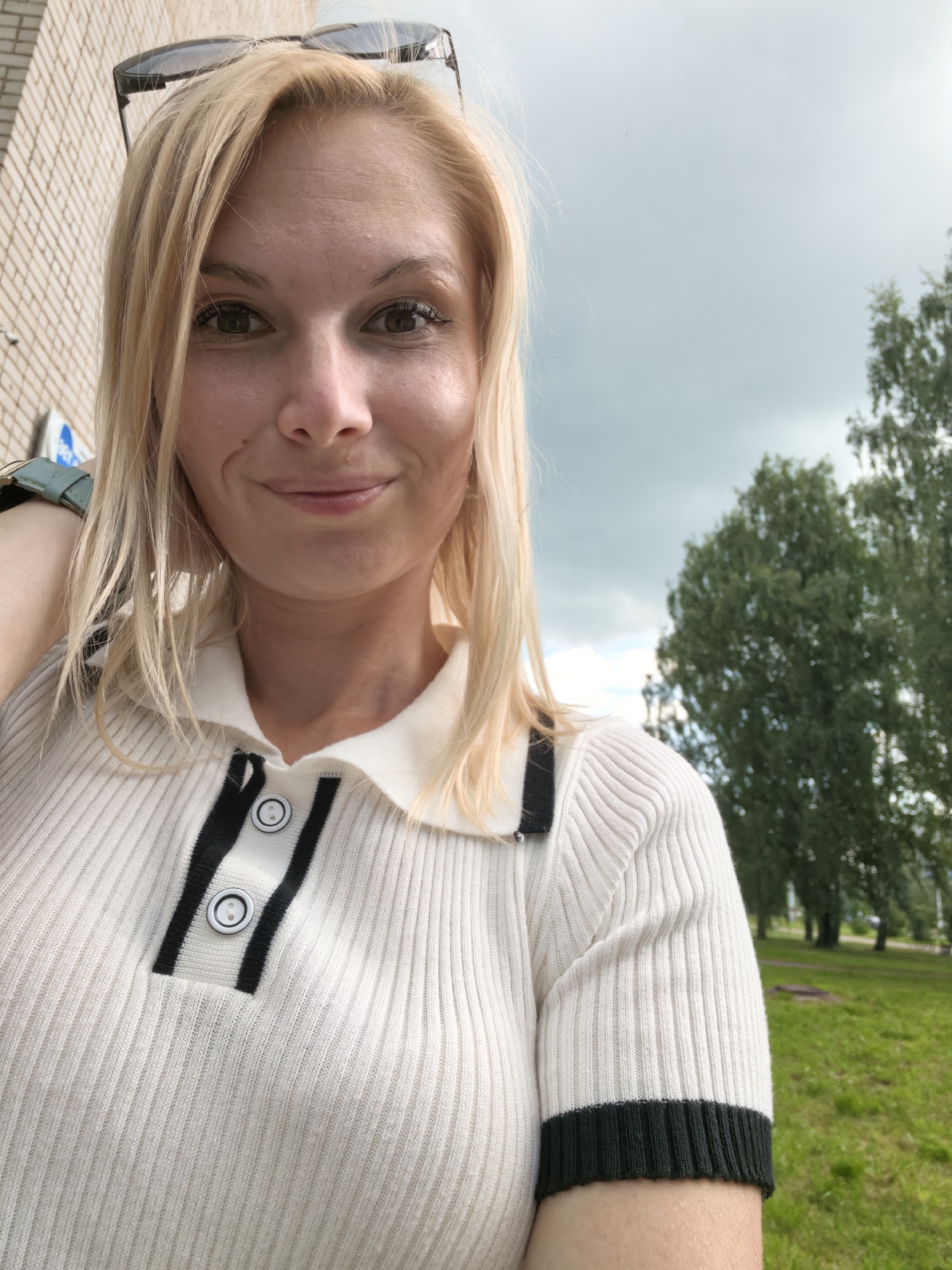 A woman with blonde hair and sunglasses on her head is taking a selfie outdoors. She is wearing a white ribbed polo shirt with black trim and buttons. The background shows a brick building on the left, a cloudy sky, and trees and grass on the right. The location is Sterlitamak, Russia.