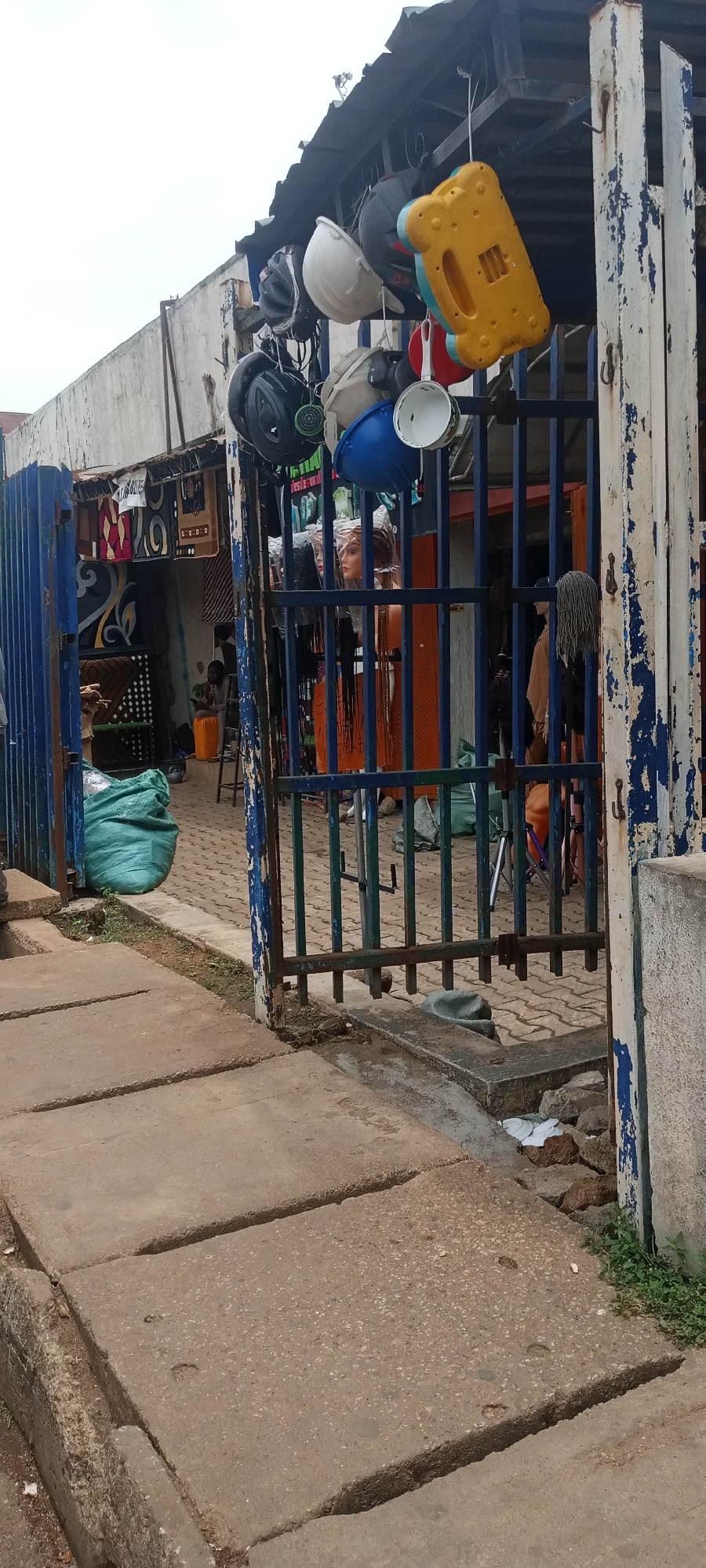 The image shows a shop entrance in Jos, Nigeria. A blue metal gate, exhibiting chipped paint, stands partially open. Items such as helmets, a blue bowl, a white saucepan, and a yellow toy are hung from the gate's top bar. Inside the shop, mannequins with braided wigs are visible. In the left corner, a large green bag rests on the ground. A seated individual is also seen inside the shop. The foreground features concrete paving stones forming a sidewalk. Litter is present near the gate. The general environment suggests an outdoor marketplace or commercial area.