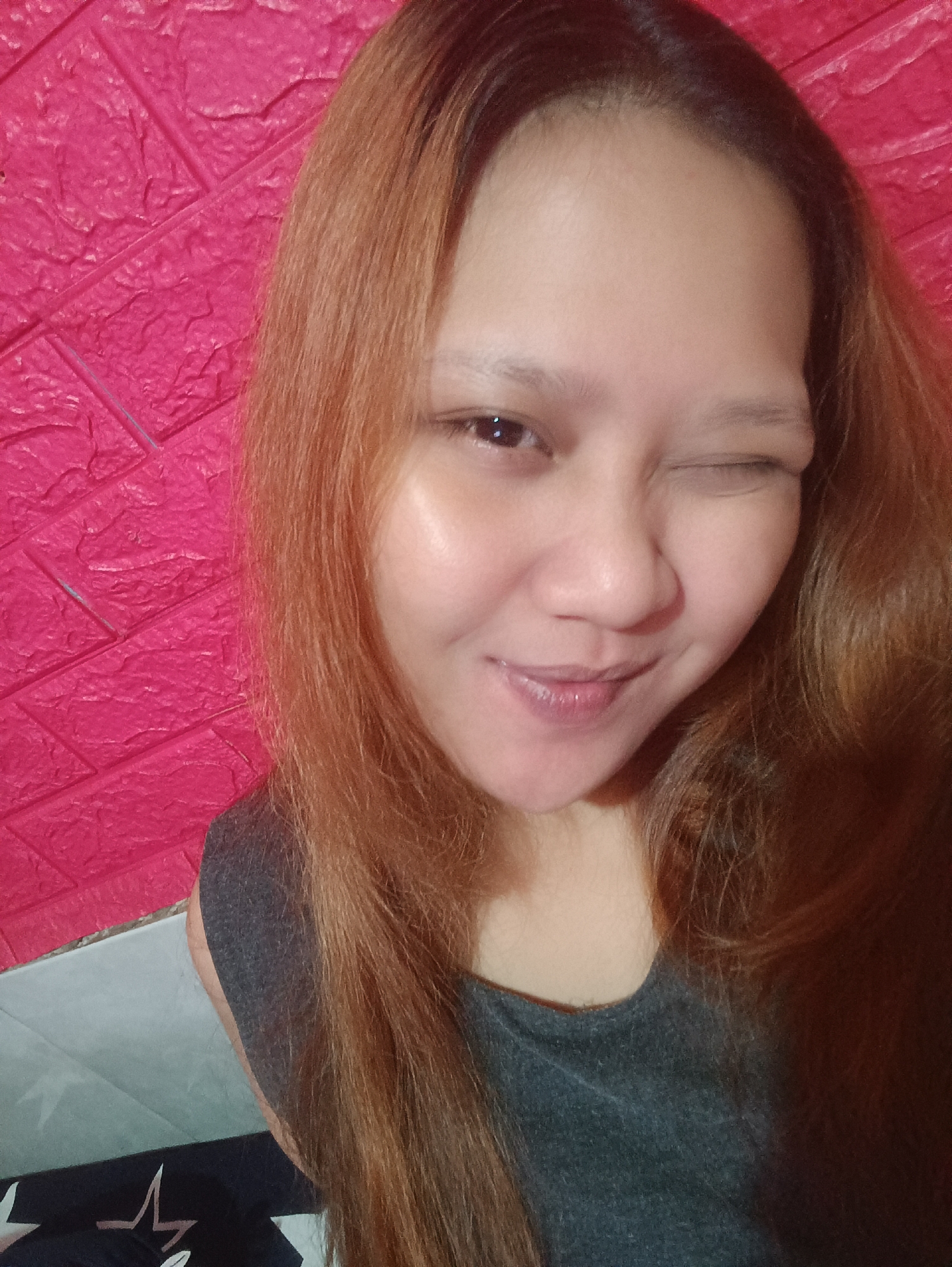 The media file is a close-up, waist-up selfie of a young woman with long, reddish-brown hair. She is looking directly at the camera and winking. Her skin appears smooth and has a dewy sheen. She is wearing a dark grey, sleeveless top.
The background consists of a textured, bright pink wall on the left, which appears to be made of foam bricks or a similar material. To the bottom right of the image, there is a hint of a grey and white tiled surface, possibly a floor or counter, and a black cloth with white star patterns.
The lighting suggests it is indoors. The overall mood of the image is playful and casual, conveyed by the wink and the slight smile. There is no visible text in the image. The location context is Depok, Indonesia.