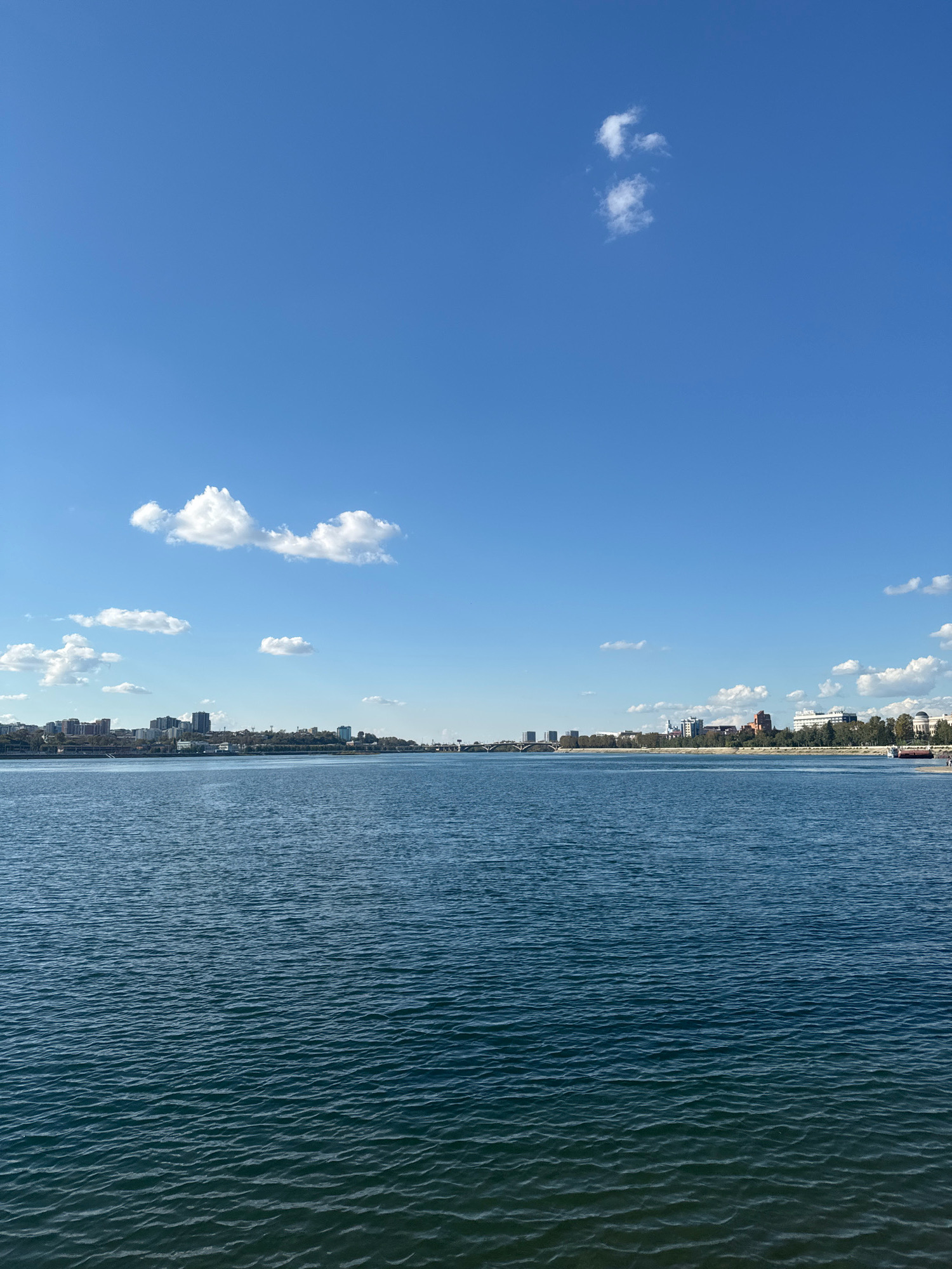 The image depicts a wide body of water, likely a river, under a clear blue sky dotted with a few fluffy white clouds. On the far bank of the water, a city skyline is visible, characterized by a mix of modern buildings and some older structures. Trees line the waterfront, and several bridges can be seen spanning the water in the distance. The water itself has a textured surface with small ripples, suggesting a gentle breeze. The scene appears to be an urban waterfront, possibly Irkutsk, Russia, given the architectural style and general ambiance. There are no visible people or distinct actions occurring in the frame. The overall impression is one of a calm and sunny day in a city situated by a substantial body of water.