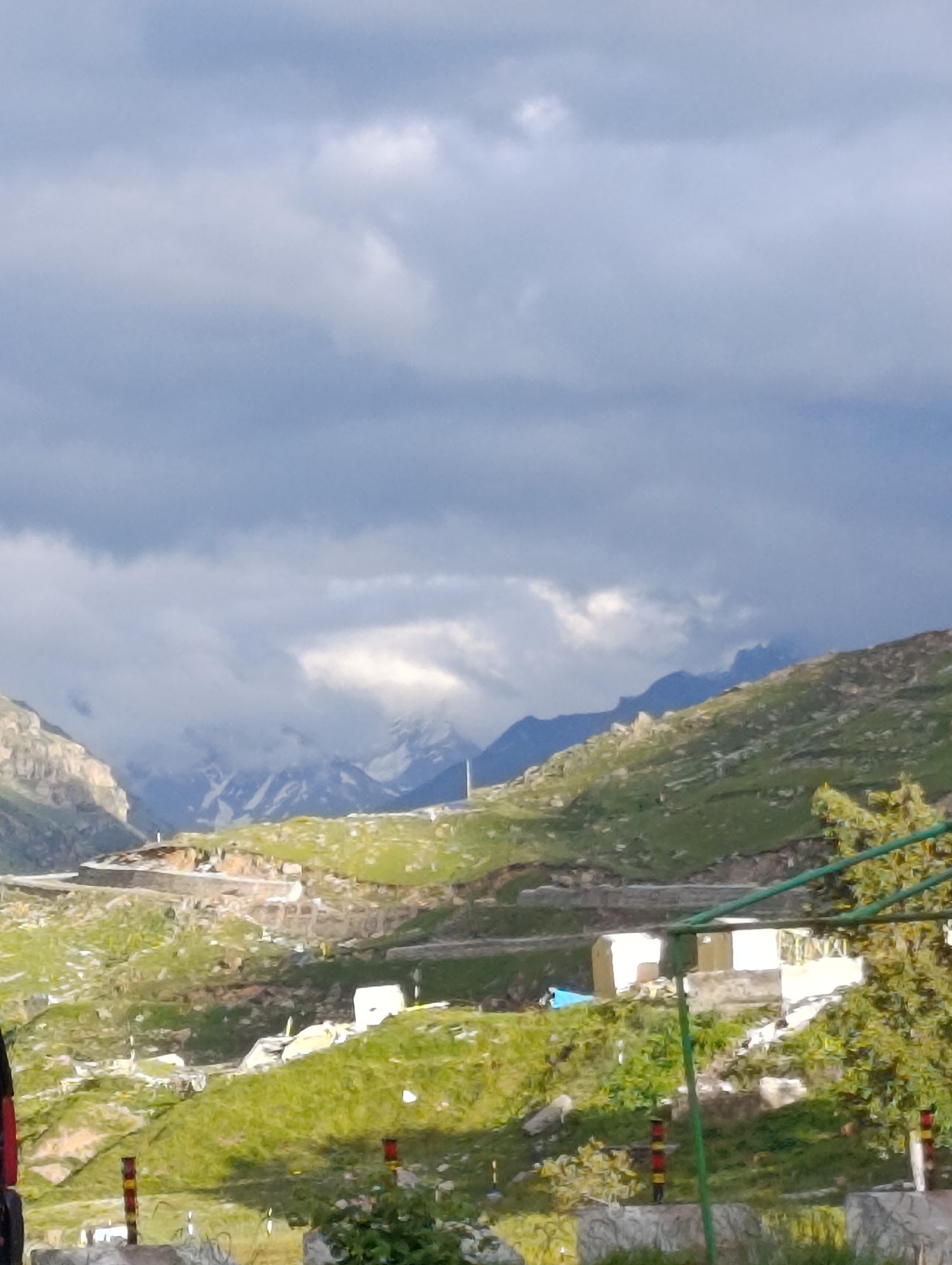 The image shows a mountainous landscape in Damphug, India, under a cloudy sky. The foreground features a dirt road with some scattered stones and small plants. In the midground, there are green, grassy hills with some rocky outcrops and a few low-lying buildings. A white van is parked on the road. In the background, majestic snow-capped mountains are visible, partially obscured by clouds. The overall impression is one of natural beauty and rugged terrain.