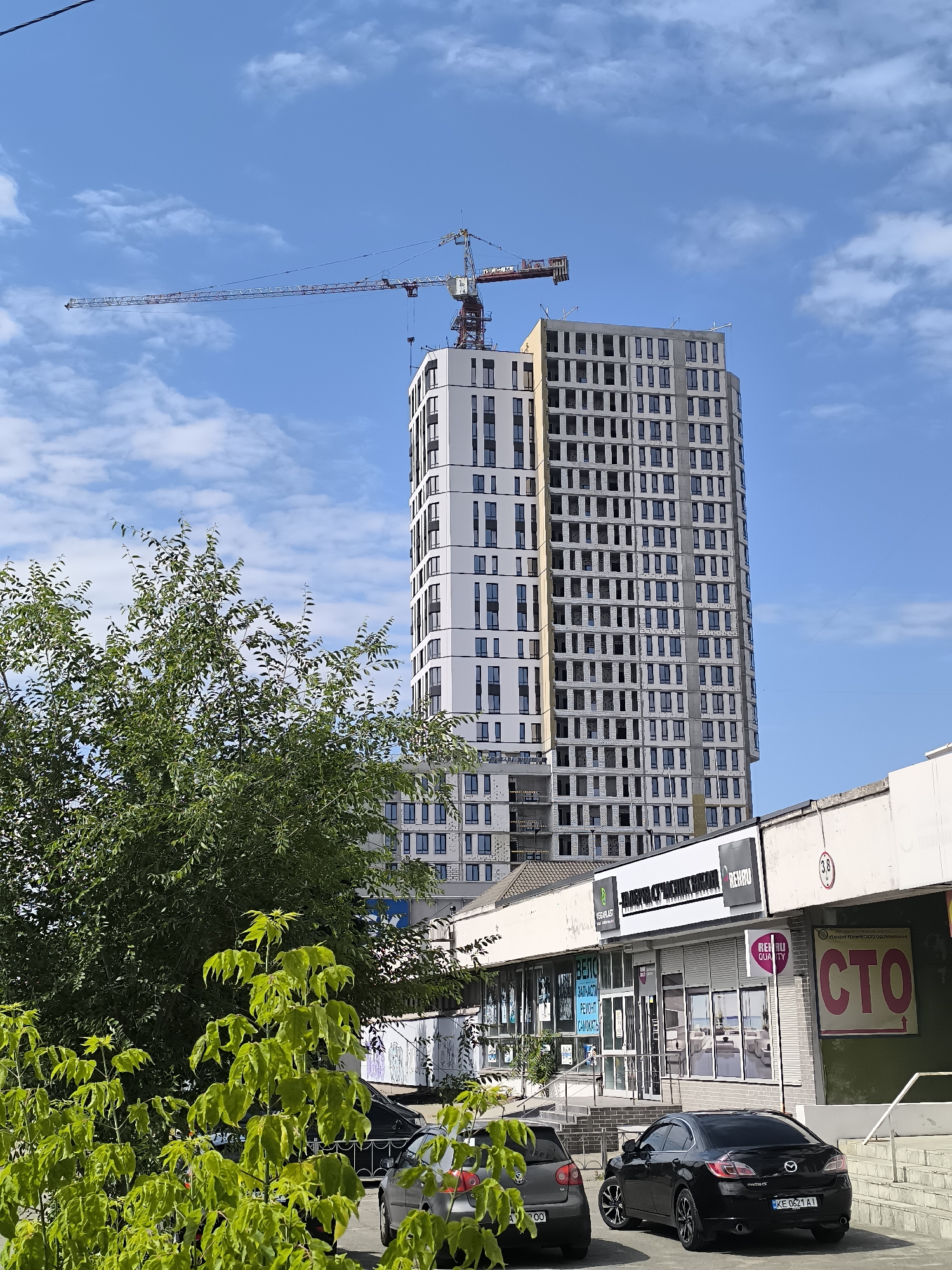 The image captures a street scene in the city of Dnipro, Ukraine, on a clear day with a bright blue sky dotted with white clouds. In the background, a tall, modern apartment building is under construction, its exterior clad in white and grey panels, with numerous windows visible but not yet installed. A large construction crane with a red boom extends over the building. In the mid-ground, a row of commercial establishments with large storefront windows is situated. Signs above the shops include "REHOU QUALITY" and "СТО," the latter indicating a vehicle service station. Green trees, one in the foreground with bright green leaves, partially obscure the view of the buildings. Several cars are parked on the street in front of the shops, including a black sedan and a grey hatchback. The architecture suggests a contemporary urban environment in Dnipro.