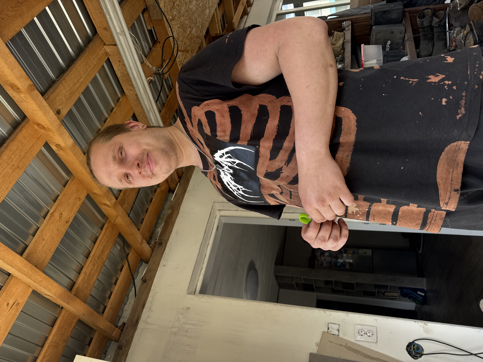 This image, captured in Chaikovsky, Russia, features a man positioned indoors in what appears to be a workshop, shed, or a room undergoing renovation. The man, a Caucasian individual with short, light-colored hair and stubble, looks directly at the camera with a calm, slight smile. He is wearing a black t-shirt adorned with a distinctive design of brown and white patterns, reminiscent of a skeletal ribcage or abstract organic shapes, possibly created using a bleach-dye technique. A silver chain is visible around his neck. In his right hand, he holds a small, bright green clip or tool.
The background reveals an unfinished or utilitarian environment. Above the man, a ceiling structure consists of exposed wooden beams supporting corrugated metal sheets, with electrical wires running along them. To his right, a cluttered storage area holds various items, including dark boots and a black plastic case with "XPB" clearly visible on its side. Below his right arm, a light-colored wall, possibly concrete or unfinished plaster, features electrical outlets (double-round pin type) and exposed wiring. An open doorway or window in this wall provides a glimpse into an adjacent room, where a round ceiling light fixture and shelves are discernible. The scene is illuminated by natural light, suggesting it is daytime.