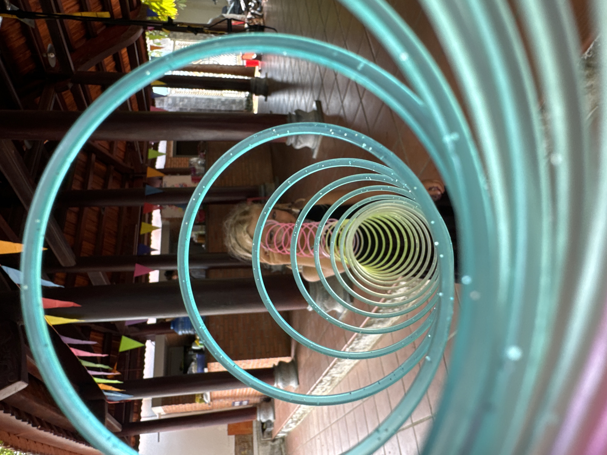 The image depicts a downward-looking perspective through a series of nested, translucent blue and yellow plastic hoops. A person wearing a pink top is located at the center, with their hair visible in the background. The view reveals an outdoor area, possibly a courtyard or patio, with wooden structures, brick walls, and tile flooring. There are colorful triangular pennants strung along a beam. The background shows glimpses of the surroundings, with building materials. The photograph was taken in Mũi Né, Vietnam.