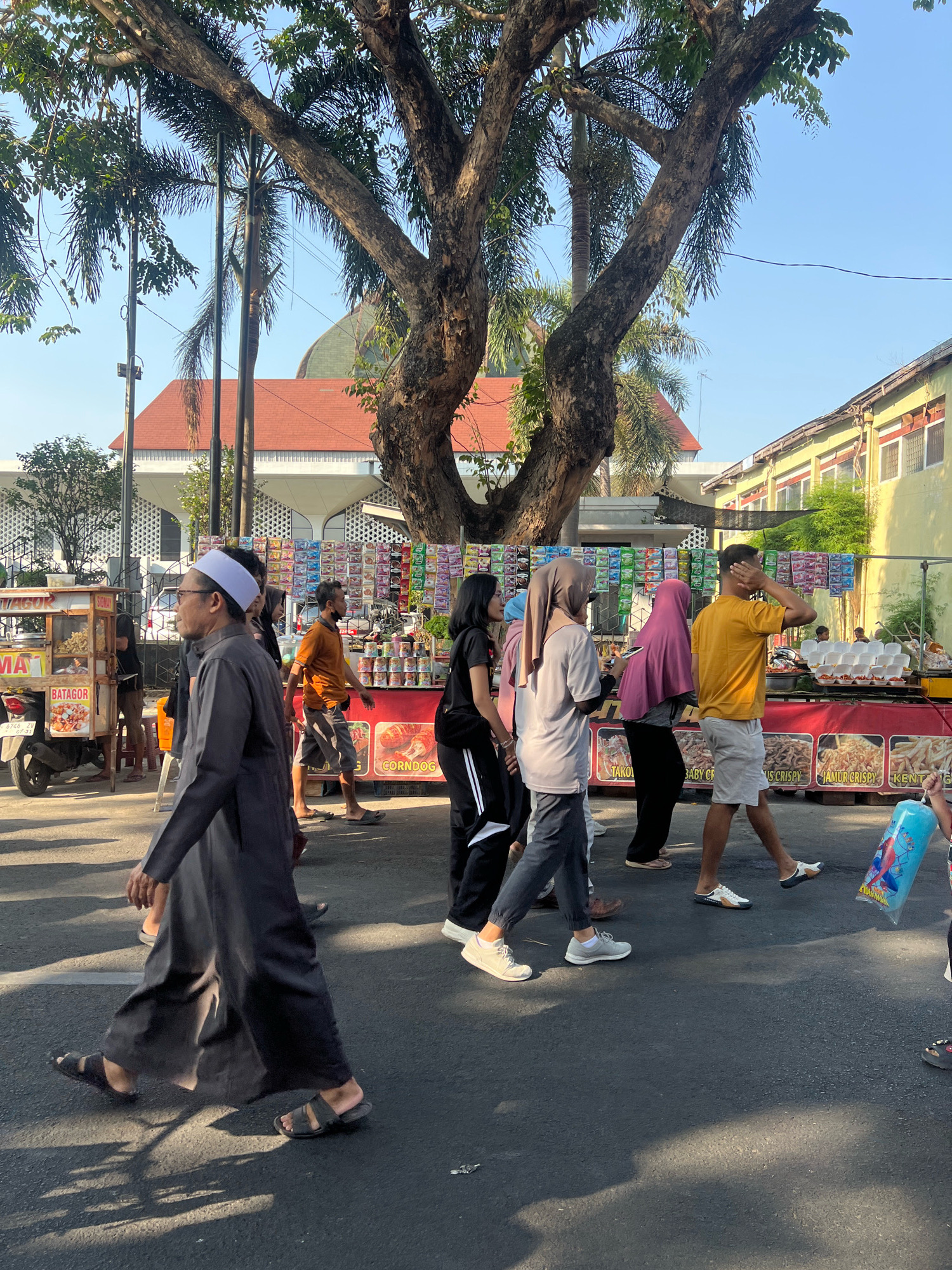 A group of people are walking on a street in Kecamatan Pati, Indonesia, on a sunny day. In the foreground, a man wearing a dark robe and white cap walks to the left. Behind him, a group of people are walking in the same direction. To the left, a food cart is parked on the side of the road. In the background, there are buildings and trees. A banner across the street displays various snacks for sale. The man in the foreground is the closest to the camera and is walking on the asphalt road, which is partially shaded by trees. The other people are walking on the same road, and the food stands are set up on the sidewalk. The scene appears to be a market or a street fair.