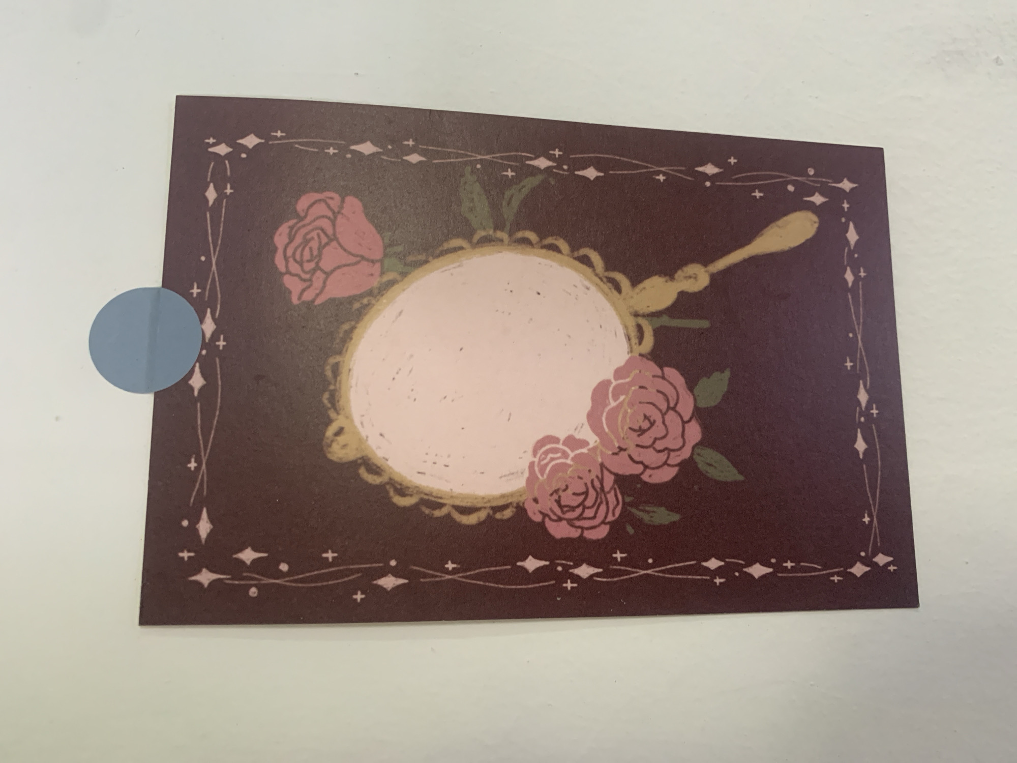 A dark purple postcard, adorned with a decorative border of pink crosses and diamonds, is attached to a white wall with a blue circular sticker. The postcard features an illustration of a vintage hand mirror with a golden handle and a scalloped edge. A pale pink oval shape represents the mirror's reflective surface. Three pink roses with green leaves are depicted, one above and to the right of the mirror, and two below and to the left. The scene is indoors, with a plain white wall as the background. The location is not specified, but the question mentions Kuala Pembuang, Indonesia, however, there is no information within the image to confirm this.