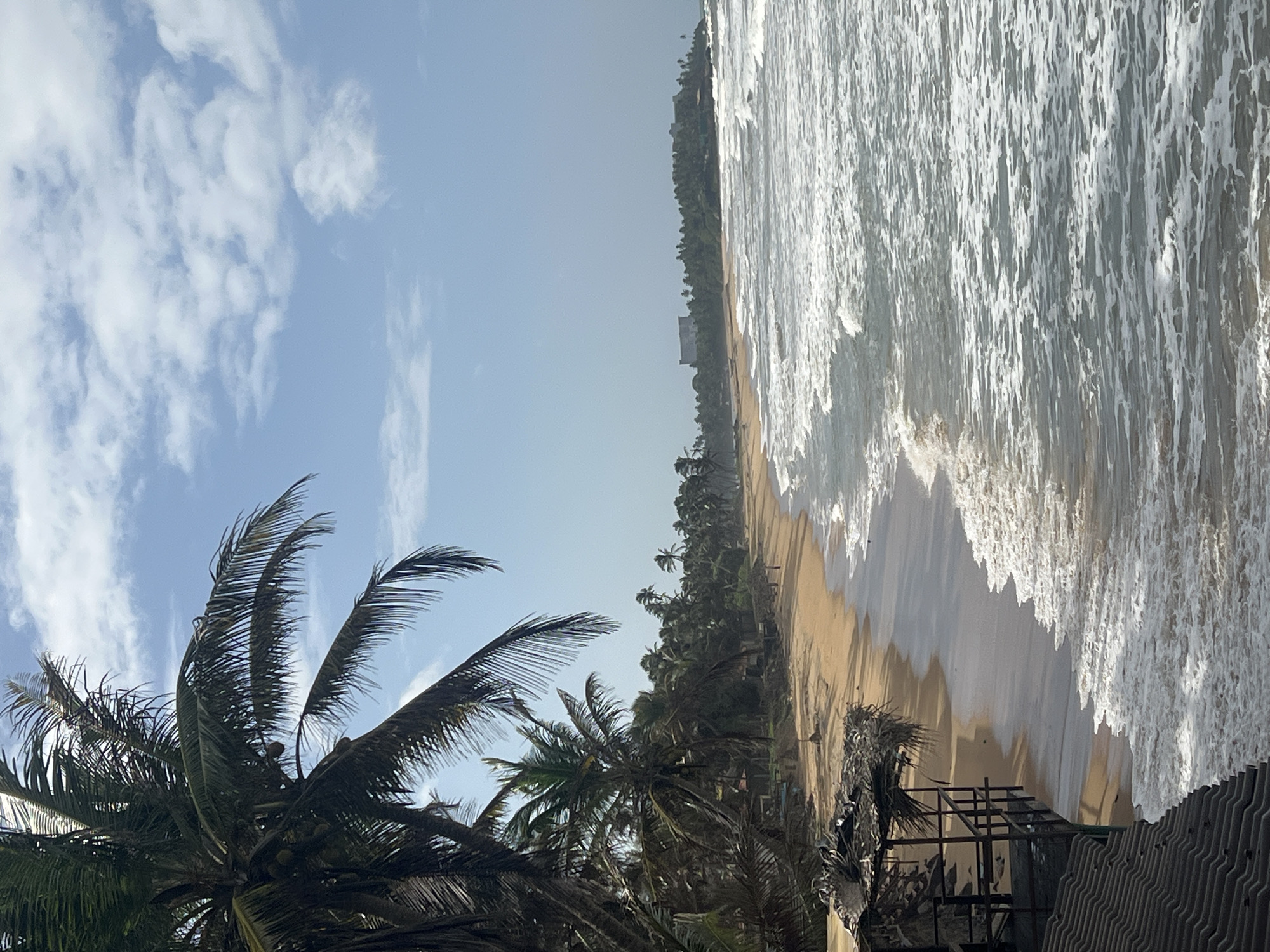 A wide-angle, vertical shot captures a tranquil beach scene in Rajgama, Sri Lanka. The foreground is dominated by a dark, textured roof, hinting at a nearby structure. To its right, a small, rustic hut with a thatched roof and open wooden platform stands near the water's edge, providing a shaded vantage point. Beyond the hut, a golden sandy beach stretches along the coastline, meeting the gentle ebb and flow of the ocean. Waves with foamy crests crash onto the shore, creating a dynamic interplay of water and sand.
A line of palm trees forms a lush backdrop, casting dappled shadows on the beach. In the distance, a cluster of buildings, possibly a resort, is visible amidst the dense greenery. The sky is a pale blue, dotted with scattered white clouds, contributing to the serene atmosphere. A few distant figures can be seen walking along the beach, adding a sense of human presence to the otherwise natural landscape. The image conveys a sense of peaceful isolation and tropical beauty.