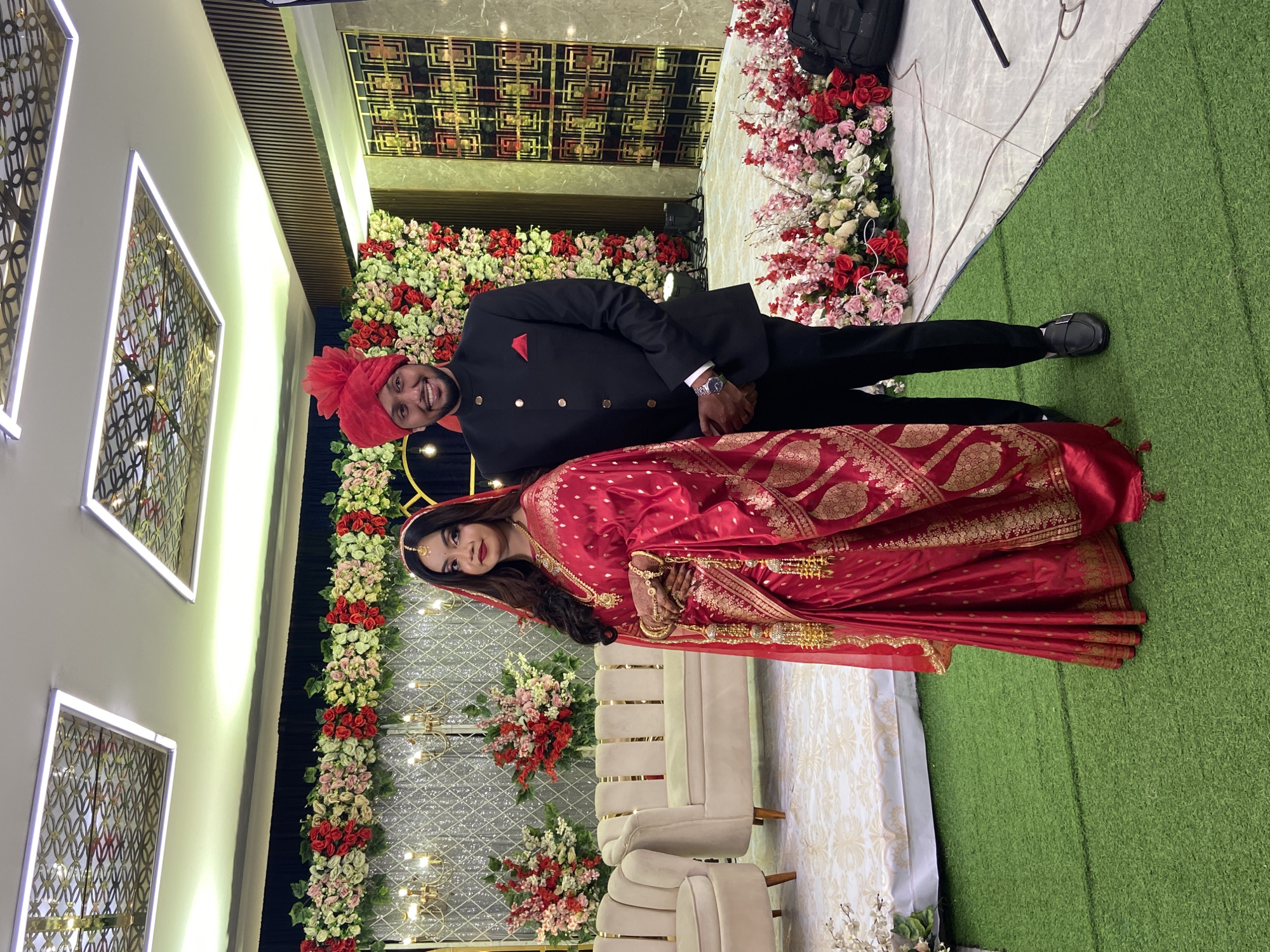 A wedding photo shows a couple in a decorated indoor location. The man wears a black suit with gold buttons, a red pocket square, and a red turban. The woman wears a red sari with gold detailing. They are standing in front of a backdrop of flowers and lights. Beige sofas are visible in the foreground. Based on the cultural attire of the couple, and the commonality of weddings being celebrated in the city with such decor, this image likely originates in Dhaka, Bangladesh.