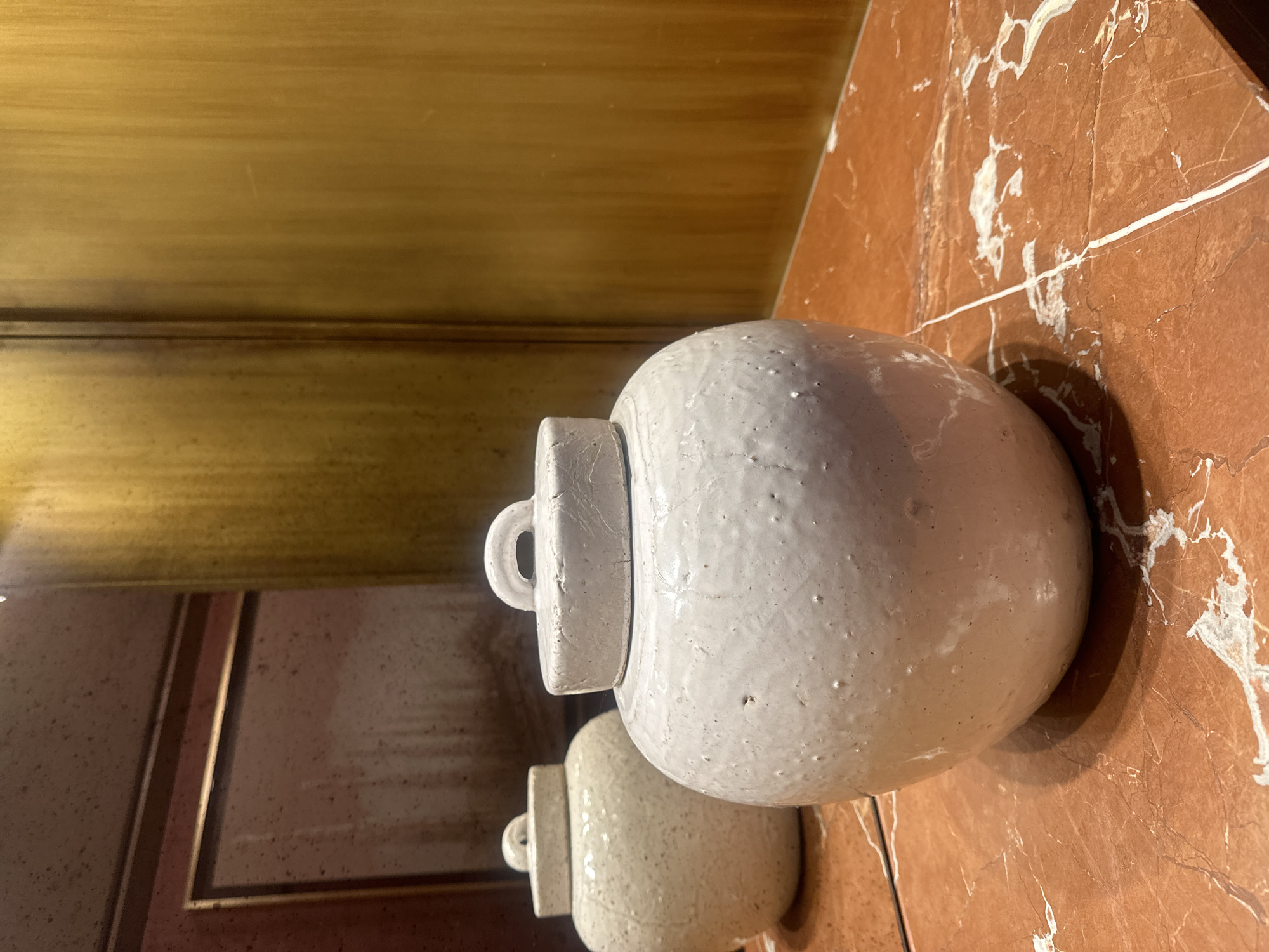 A high-angle indoor shot features two textured, off-white ceramic jars with lids, situated on a reddish-brown marble surface with white veining. The larger jar, closer to the camera, has a visible lid with a loop handle. The second jar is positioned behind and slightly below, partially obscured by the first. Behind the jars and to the left, a gold-toned cabinet or panel is visible. A framed reflection shows a portion of a matching ceramic jar. No people are visible. The location is not specified in the image.