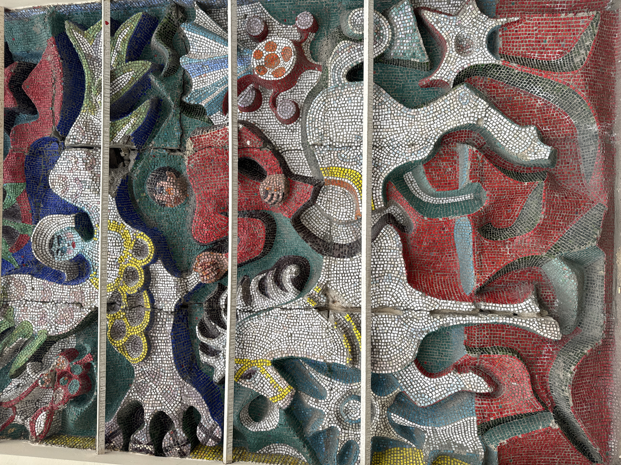 The image shows a colorful mosaic mural in the city of Unknown, Unknown. The mural is divided into four vertical panels, each separated by a white structural support. The mosaic is composed of small, square tiles in a variety of colors, including red, white, blue, green, and yellow. The design appears abstract, with swirling shapes and patterns that resemble organic forms and celestial bodies. The tiles have been meticulously arranged to create a textured and three-dimensional effect. The overall impression is one of artistic expression and craftsmanship.