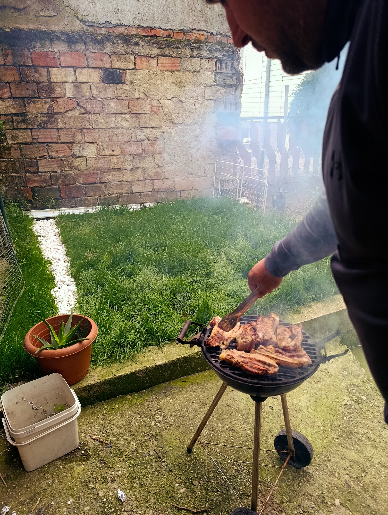 A man grills meat on a small charcoal grill in an outdoor setting. The grill, supported by three legs with two wheels, sits on a concrete patio. The man, wearing a dark gray sweatshirt, uses tongs to turn the meat. Smoke rises from the grill. Behind the grill, there is a patch of grass separated from the patio by a concrete border, with a line of white stones running along the border. To the left, a terracotta pot containing a plant and a white plastic bucket sit on the patio. A brick wall stands in the background. In the distance, beyond a wire fence and some greenery, a house with light-colored siding can be seen. The scene is in Kardzhali, Bulgaria.