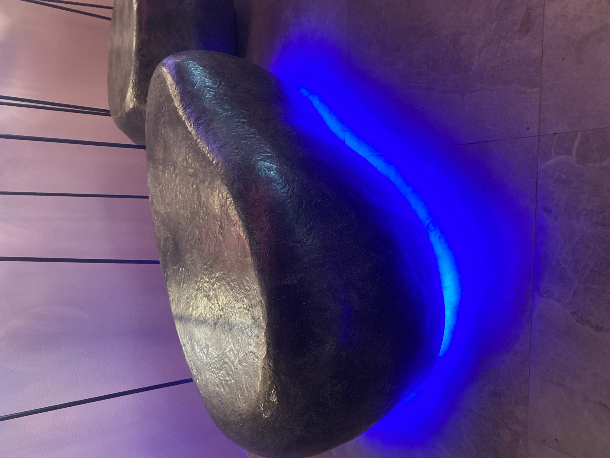 A dark, irregularly shaped bench, resembling a large, smooth stone, sits on a tiled floor. The bench is illuminated from below by bright blue LED lights, creating a vibrant glow that reflects onto the floor and the bench's underside. The bench has a textured, metallic-looking surface. In the background, a light purple wall is visible, with thin, dark vertical lines extending upwards. Another similar bench is partially visible to the right. The scene appears to be indoors, possibly in a modern public space or art installation. There are no people visible in the image. The lighting suggests it may be evening or a dimly lit environment. The flooring consists of large, light-colored tiles with a marble-like pattern. The overall aesthetic is contemporary and atmospheric. The location is Berdsk, Russia.