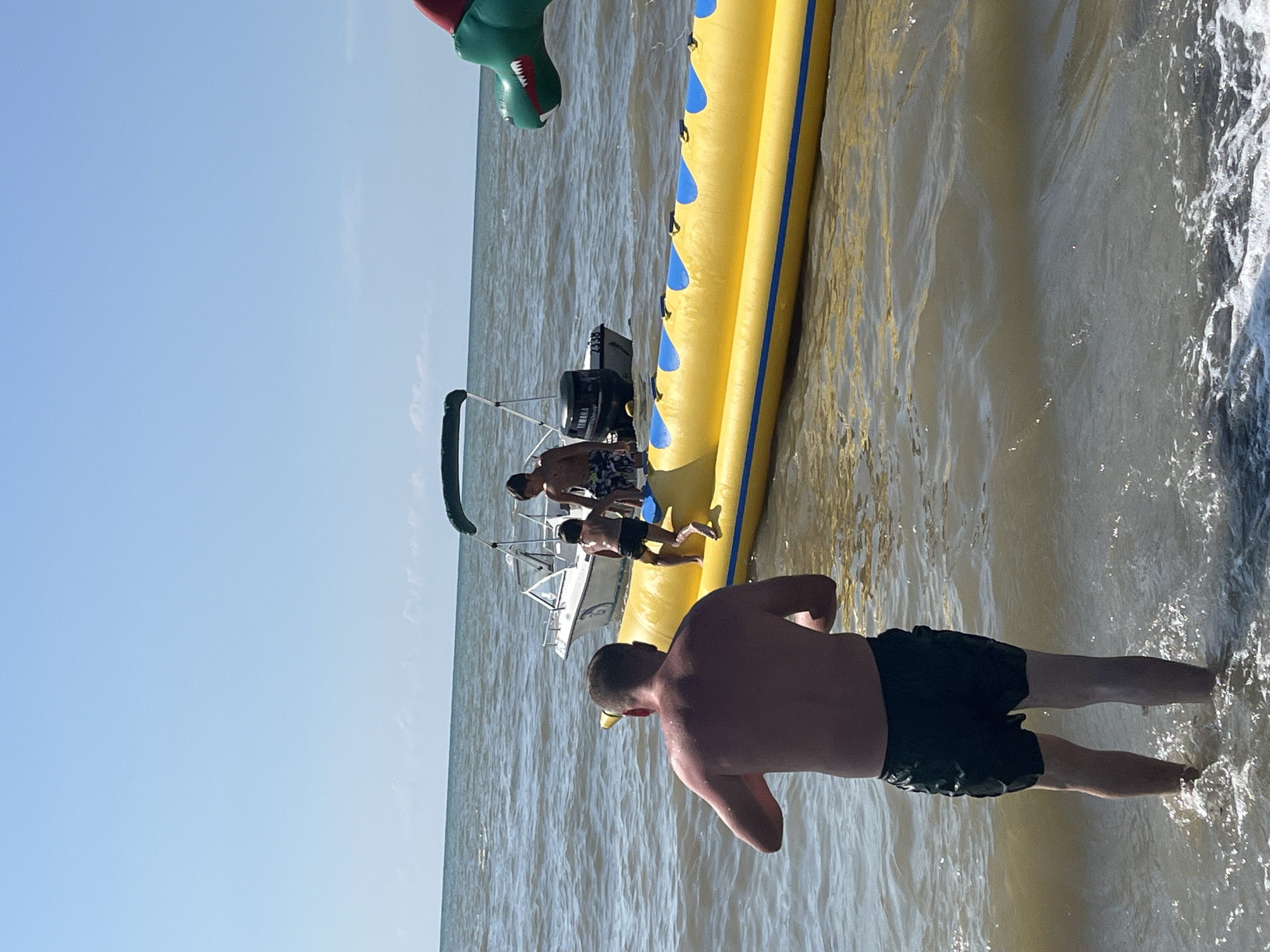 An aerial view shows a man in black swim trunks standing in shallow water at a beach. He is facing away from the camera, towards a yellow inflatable tube with blue accents, which is floating in the water. Two young boys are on the inflatable tube. A white motorboat is docked behind the tube. In the background, the sea stretches to the horizon under a clear blue sky. To the right, the green head of an inflatable dragon toy is visible in the water. The scene depicts a recreational activity at the beach of Ureki, Georgia, a coastal town known for its magnetic sand beaches.