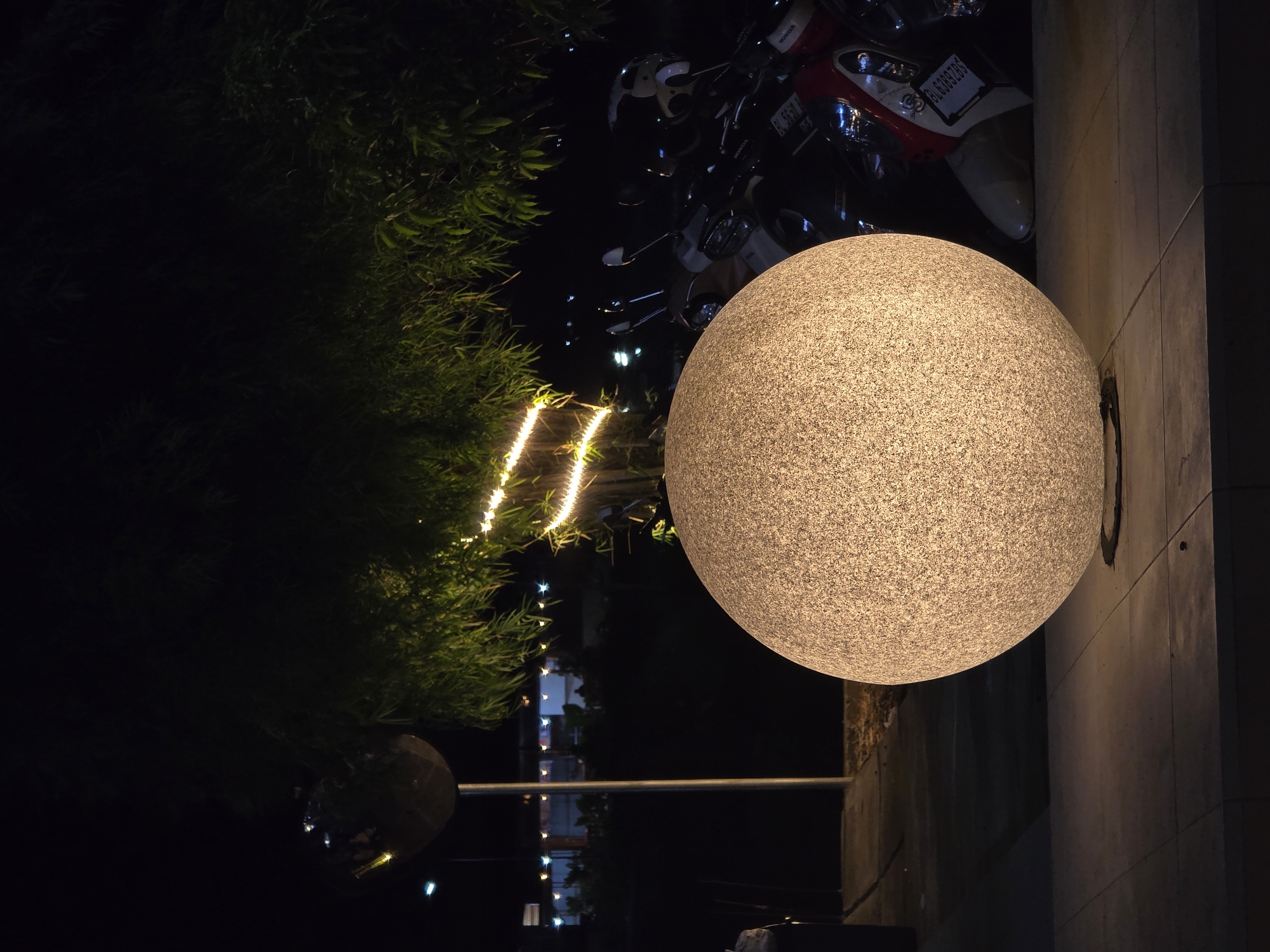 In the city of Uleeue, Indonesia, an outdoor scene at night is captured. A large, illuminated spherical lamp sits prominently in the foreground, its textured surface glowing with a warm, white light. It rests on a tiled surface. Behind the lamp, a row of parked motorcycles is visible, their lights and reflections glinting in the darkness. A tree with dense foliage extends from the upper portion of the image, adorned with a string of small, bright lights that create a festive atmosphere. In the background, distant buildings and lights suggest the presence of an urban or commercial area. A tall, slender pole with a reflective sphere on top stands to the left of the frame, further adding to the nighttime ambiance. The overall impression is one of a well-lit public space, likely a plaza or walkway, designed for evening enjoyment.
