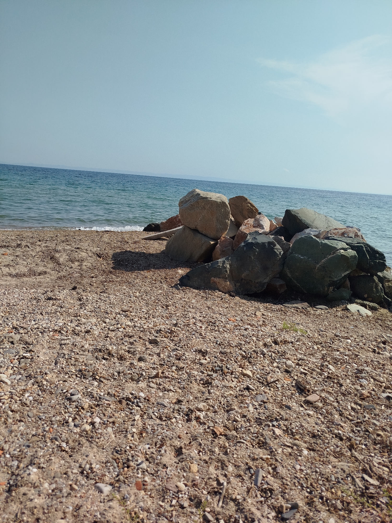 A close-up, low-angle shot captures a rocky beach with the Aegean Sea in the background. The foreground is dominated by a dense, grainy mixture of sand and small pebbles in shades of brown, grey, and white. A cluster of large, irregular rocks, varying in color from light tan to dark grey and hints of green, are piled on the left side of the frame, extending towards the water's edge.
The middle ground features the transition from the pebble-strewn beach to a strip of darker, wet sand where gentle waves are washing ashore, leaving a white foam line. The water is a clear, bright blue, reflecting the sky. In the distance, a hazy, light blue horizon line suggests landmasses or mountains. The sky is a pale, clear blue, with only a faint, wispy cloud visible on the right side, high up. The lighting suggests a bright, sunny day, with shadows cast by the rocks. The scene is devoid of people or any manufactured objects. The location is a natural beach environment within Sithonia Municipal Unit, Greece.