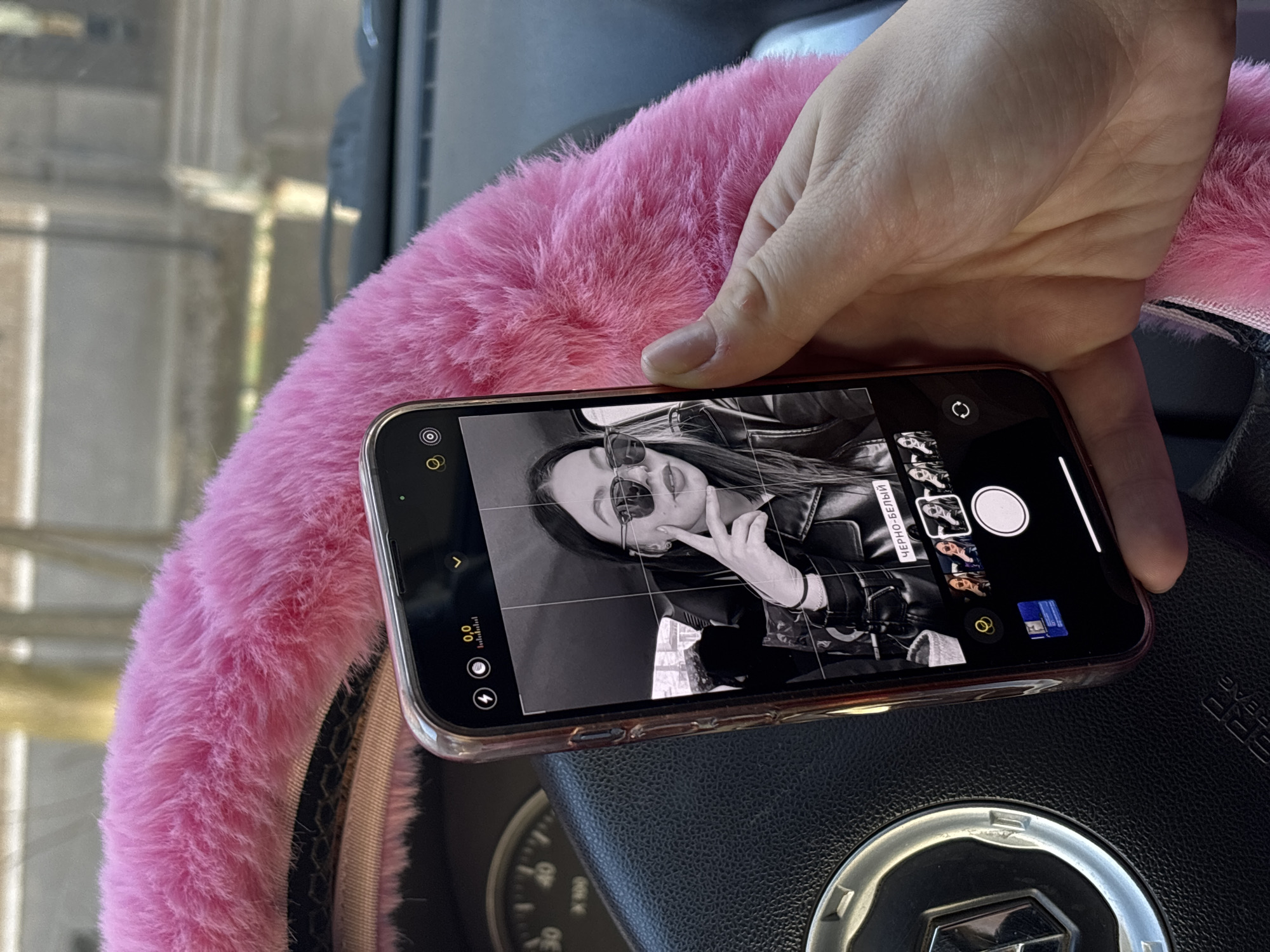 A person is holding a smartphone over a car steering wheel. The smartphone's screen displays a black and white photo of a person wearing sunglasses and a leather jacket. The steering wheel has a pink, fuzzy cover. The car's dashboard is partially visible, and the setting appears to be inside a car, potentially in a city as indicated by the presence of buildings and the vehicle's dashboard. The steering wheel logo indicates the car is a Renault, and it is possible to infer, from this image, that it was taken in Perm, Russia.