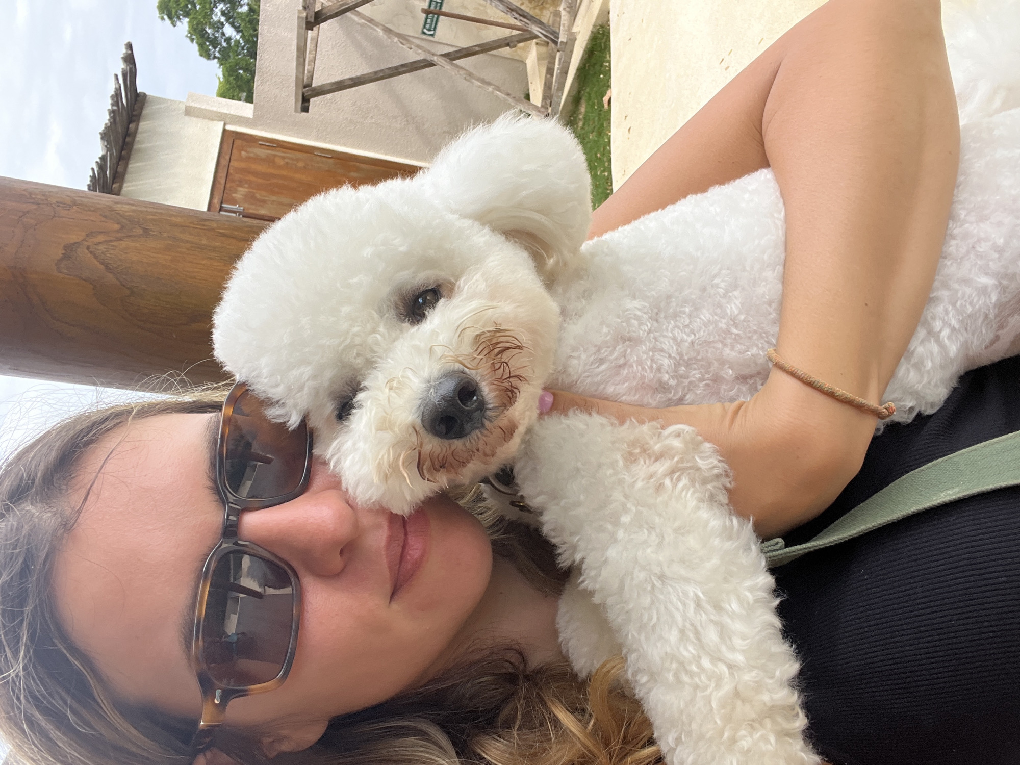 A close-up, outdoor, selfie-style photograph shows a fair-skinned woman with blonde hair and sunglasses holding a white poodle. The woman is wearing a black top. The poodle is positioned in front of the woman's face, obscuring her mouth and nose. The background is out of focus, but it appears to be a rustic, possibly tropical setting, with wooden structures and vegetation visible. The image was taken in Kediri, Indonesia.