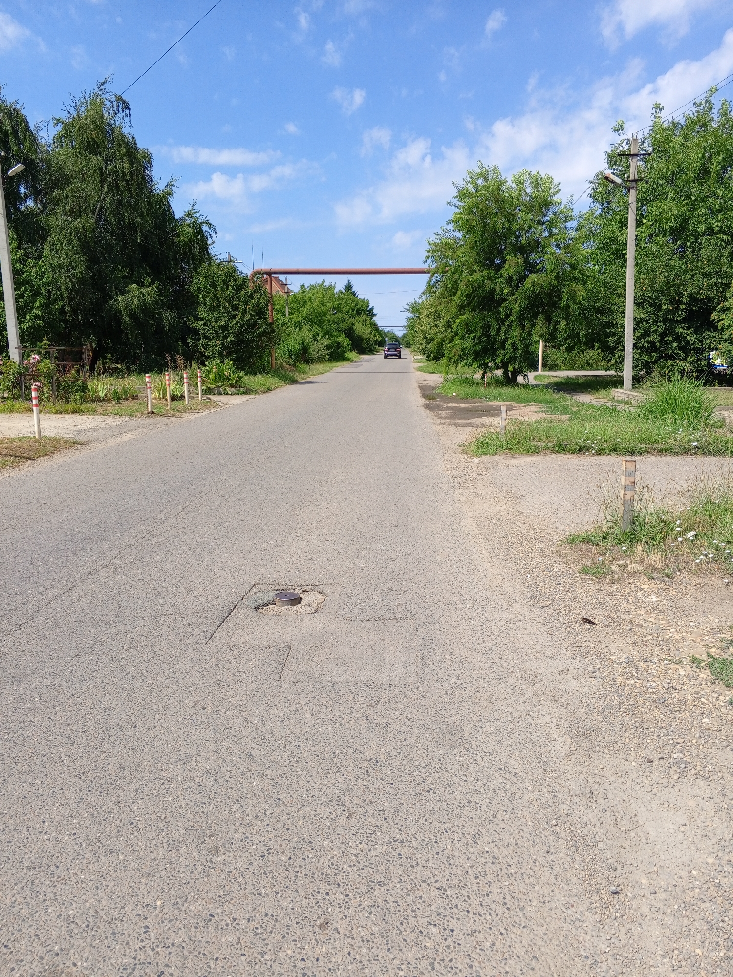 A road runs through a rural or suburban area of Lenina, Russia. The road is paved with asphalt and has a few cracks and manhole covers visible. On either side of the road are trees and vegetation. There are power lines and poles visible along the road. In the distance, a dark-colored car is driving away from the camera. The sky is blue with some white clouds. There are several small, white posts with red and white stripes marking the side of the road. There is also a large red pipe that crosses over the road in the distance.