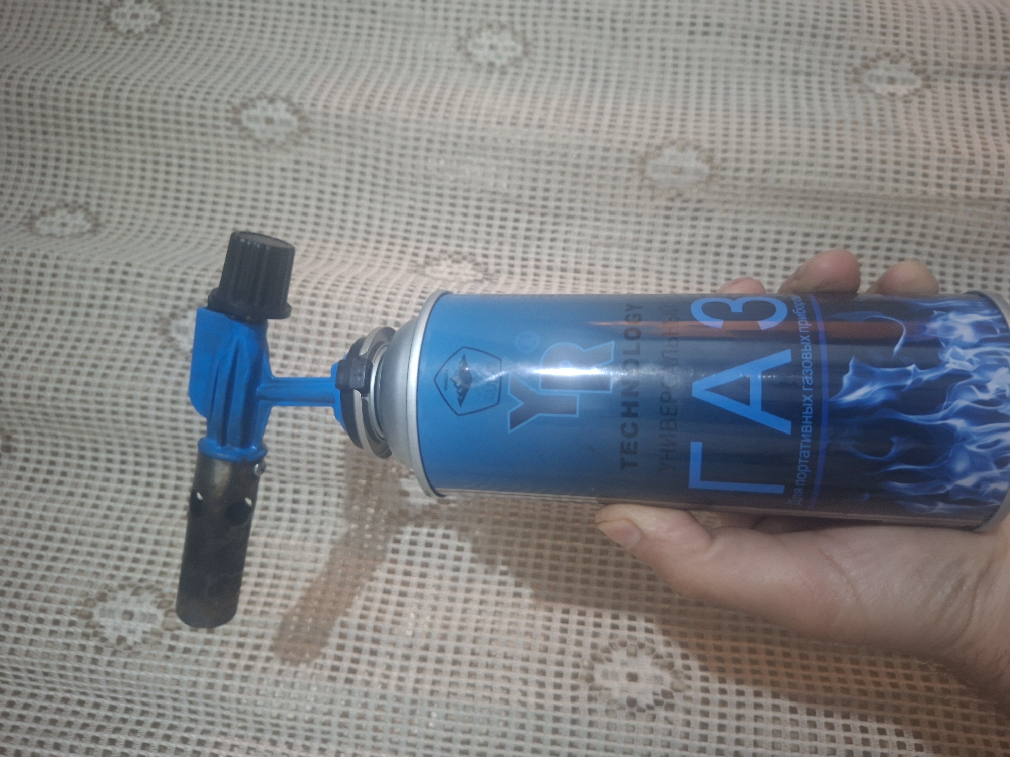 A hand holds a blue gas canister with a blue torch nozzle attached. The can has white lettering in Russian, and an image of blue flames. The background is a light-colored, patterned surface. The location is Unknown, Unknown.