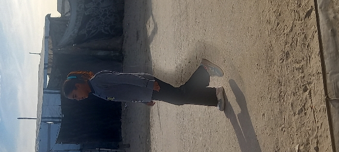 A person stands on a sandy, neutral-toned ground next to makeshift dwellings made of tarp or similar material under a cloudy sky. The individual wears a long-sleeved gray shirt and dark pants, and the shadow indicates the sun is at an angle. The location appears to be a camp or temporary settlement in a dusty environment.