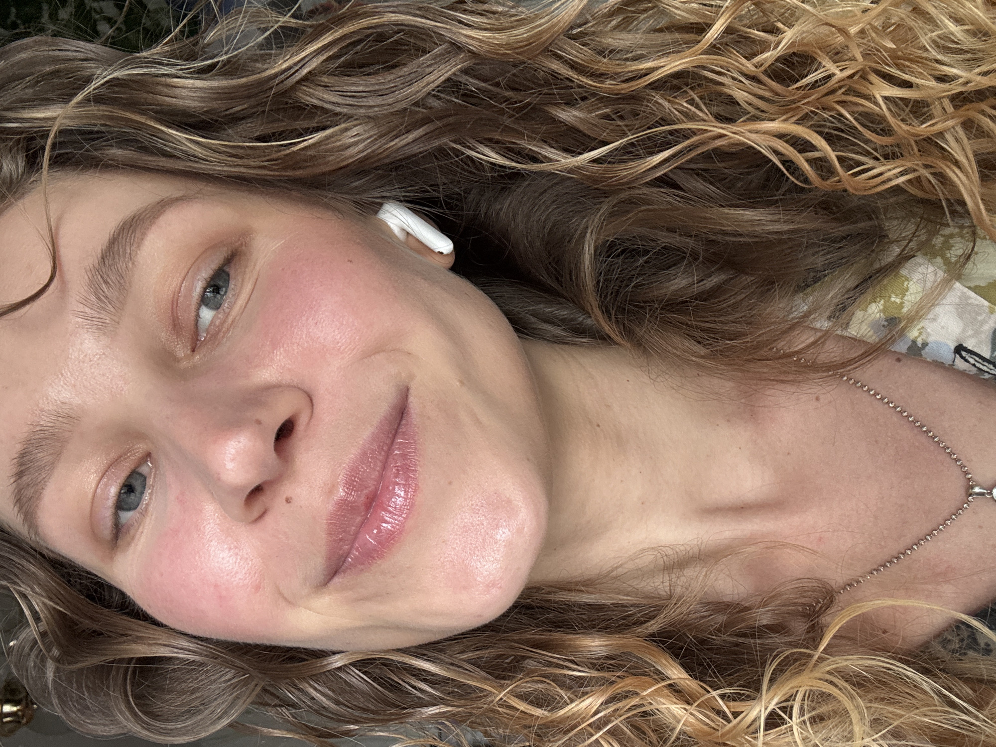 A close-up portrait of a woman with curly, light brown and blonde hair. She has light skin, blue eyes, and is wearing a white earbud in her left ear. A silver necklace is visible on her neck. There is no evidence to suggest the location is in Ketoshi, Russia. The environment is not distinctly identifiable, suggesting the focus is on the individual.