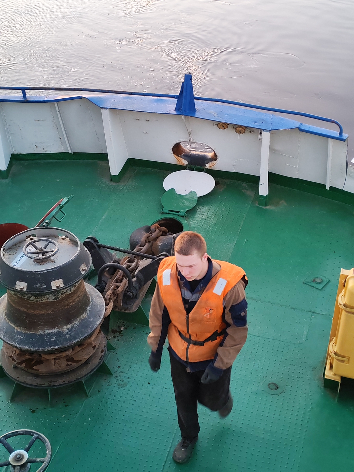 A high-angle shot shows a man in an orange life vest walking on the green deck of a boat. The deck is dotted with small, raised circles. The boat has a blue and white railing on the right side. A rusted black winch with a wheel sits on the left. Behind the man are a black anchor and chain. A yellow object is partially visible in the lower right corner. The location is likely Сигнал Снт, Russia. The water can be seen beyond the boat's railing.