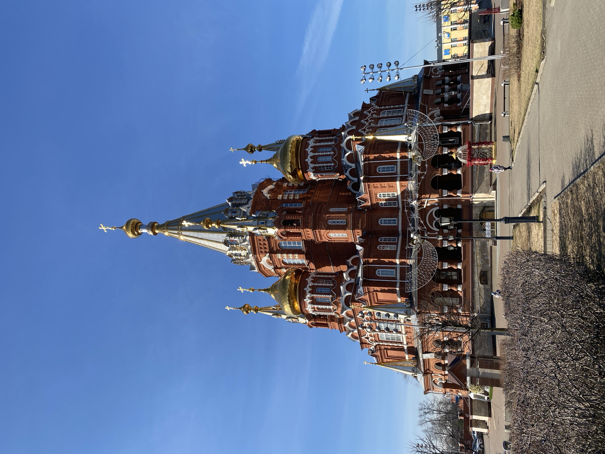 The image shows the architectural details of the Cathedral of Archangel Michael in Izhevsk, Russia, against a clear blue sky. The red brick cathedral features white window trim, gold domes topped with crosses, and a tall spire. A paved walkway runs alongside the building, with some trees and bushes. Decorations, possibly for a holiday, are visible along the side of the building.