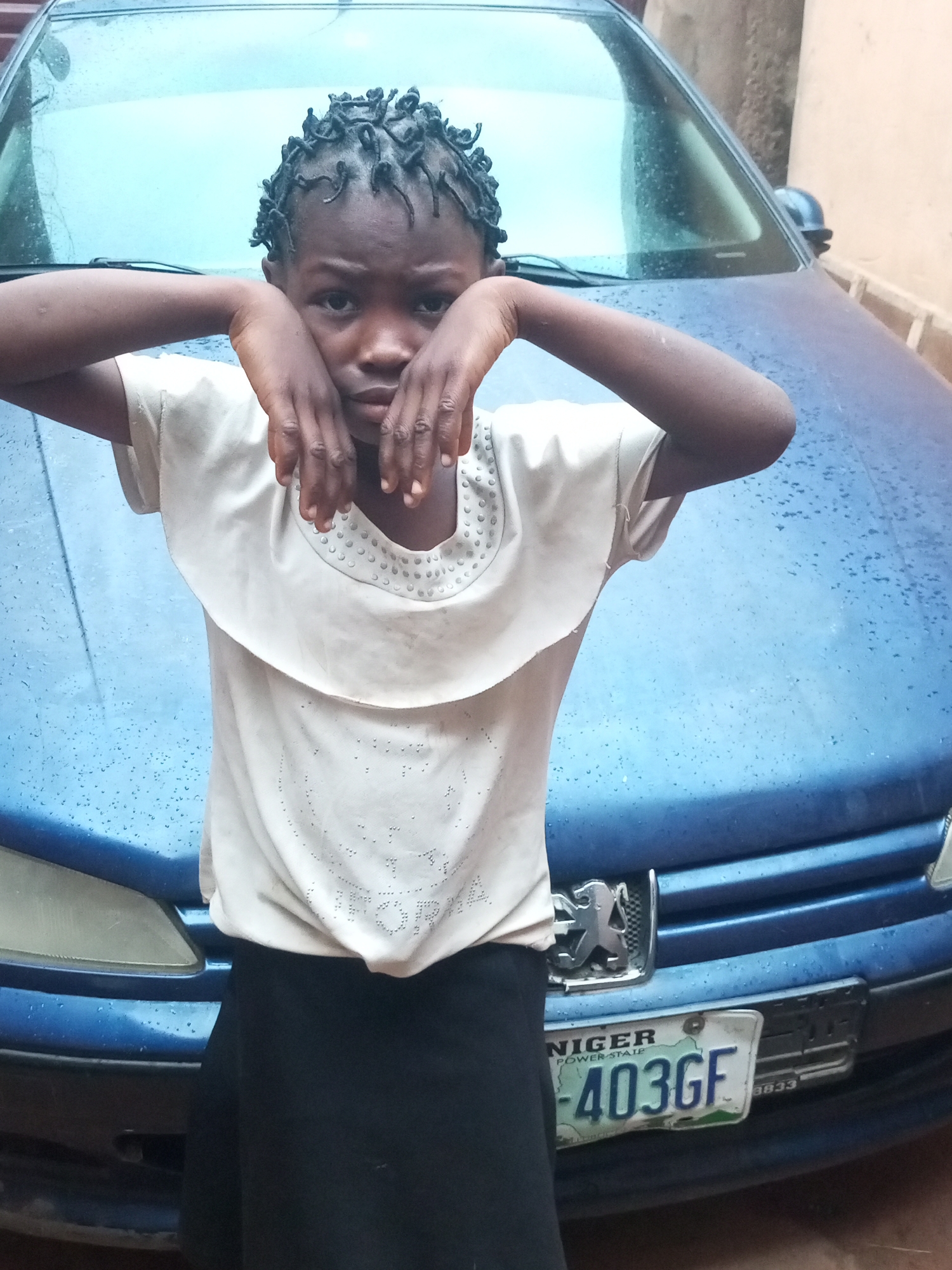 A young girl with dark skin and braided hair stands in front of a blue car. Her hands are raised to her face, obscuring her mouth. She is wearing a light-colored, ruffled shirt and a black skirt. The car's license plate, visible in the lower right portion of the image, reads "NIGER POWER STATE -403GF". The car's hood is wet, indicating recent rain. The background shows a brick wall. The image appears to be taken outdoors. The license plate suggests the location is Gombe State, Nigeria.
