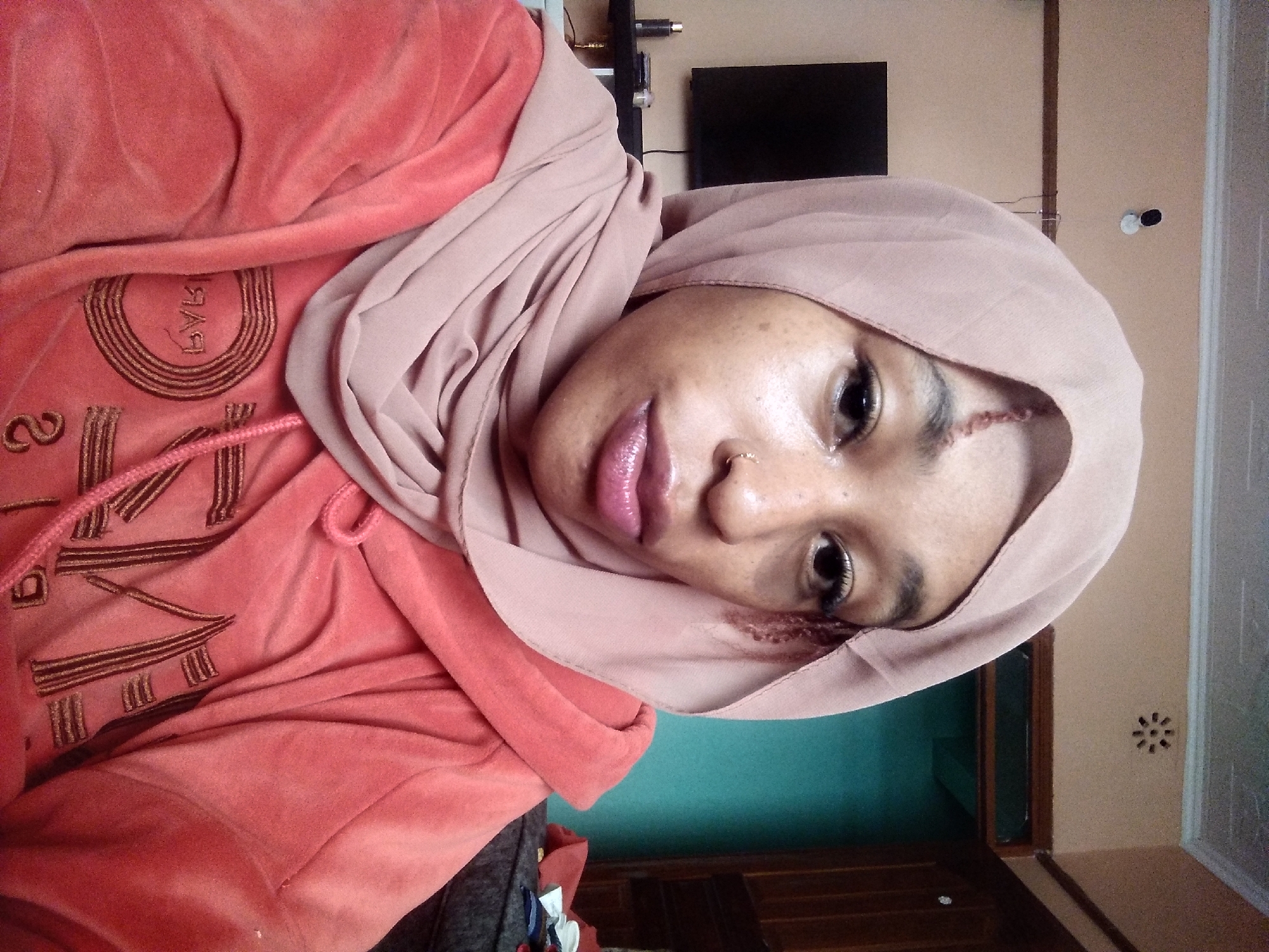 A medium close-up selfie shows a young Black woman wearing a light pink hijab and an orange velour hooded sweatshirt. She has a scar on her forehead and a nose ring. Her makeup includes eyeliner and lip gloss. The background appears to be an indoor residential setting, with a wall, ceiling, and a television visible. The room has a warm, earthy tone, with a hint of turquoise on one wall and wooden accents.