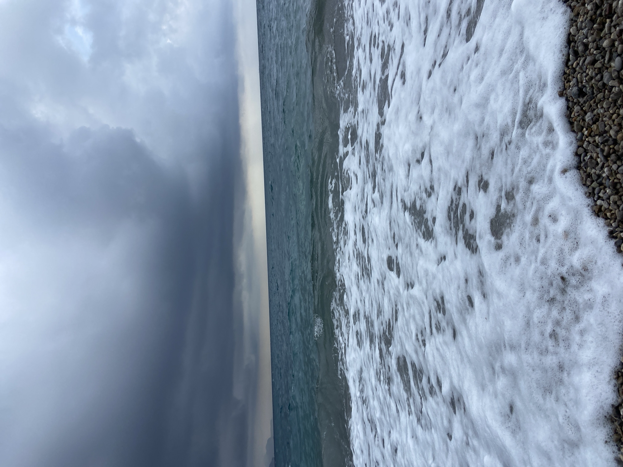 This image captures a coastal scene, likely along the Adriatic Sea in Prijevor, Montenegro, under an overcast sky. The photograph is oriented vertically, with the horizon line running down the center of the frame, separating the sky on the left from the sea and shoreline on the right.
The sky, filling the left half of the image, is dominated by heavy, dark grey clouds, suggesting stormy or very cloudy weather. There are some lighter patches visible within the cloud cover, indicating variations in thickness or glimpses of brighter sky beyond.
The sea occupies the middle band of the image, appearing a deep, turbulent blue-green. Gentle waves are visible, causing ripples and subtle movements across the surface of the water.
The right side of the image features the shoreline, where the sea meets a pebble beach. Thick white sea foam, likely from breaking waves, washes over the small, rounded grey and brown pebbles. The foam creates a textured, frothy edge where the water meets the land.
There are no people or man-made objects visible in the frame. The overall impression is one of natural beauty and a somewhat somber atmosphere due to the dark clouds. The time of day appears to be daylight, but the heavy cloud cover creates a muted light. There is no visible text.