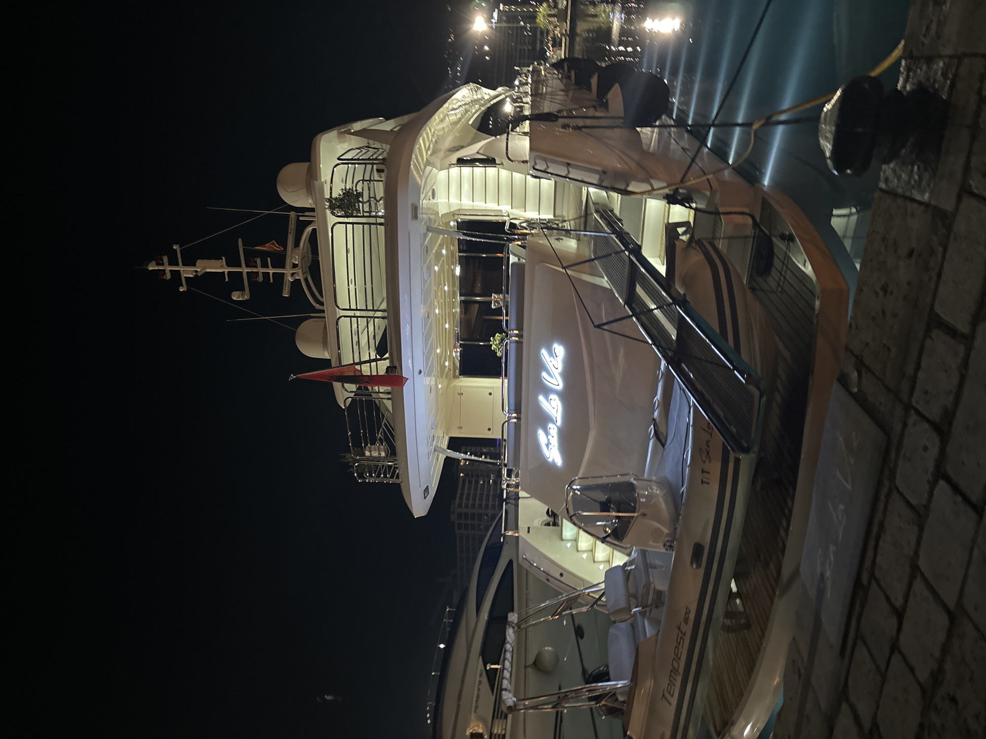 A brightly lit yacht is docked in Budva, Montenegro at night. The yacht has multiple decks and features illuminated lettering that reads "Cela Vie" on its hull. The name "Tempest 600" is also visible. The vessel is moored alongside a stone quay. Street lights are visible in the background, reflecting off the water's surface. The night sky is dark. No people are clearly visible in the immediate foreground. The scene focuses on the yacht and its illuminated features within the context of a marina or harbor setting.