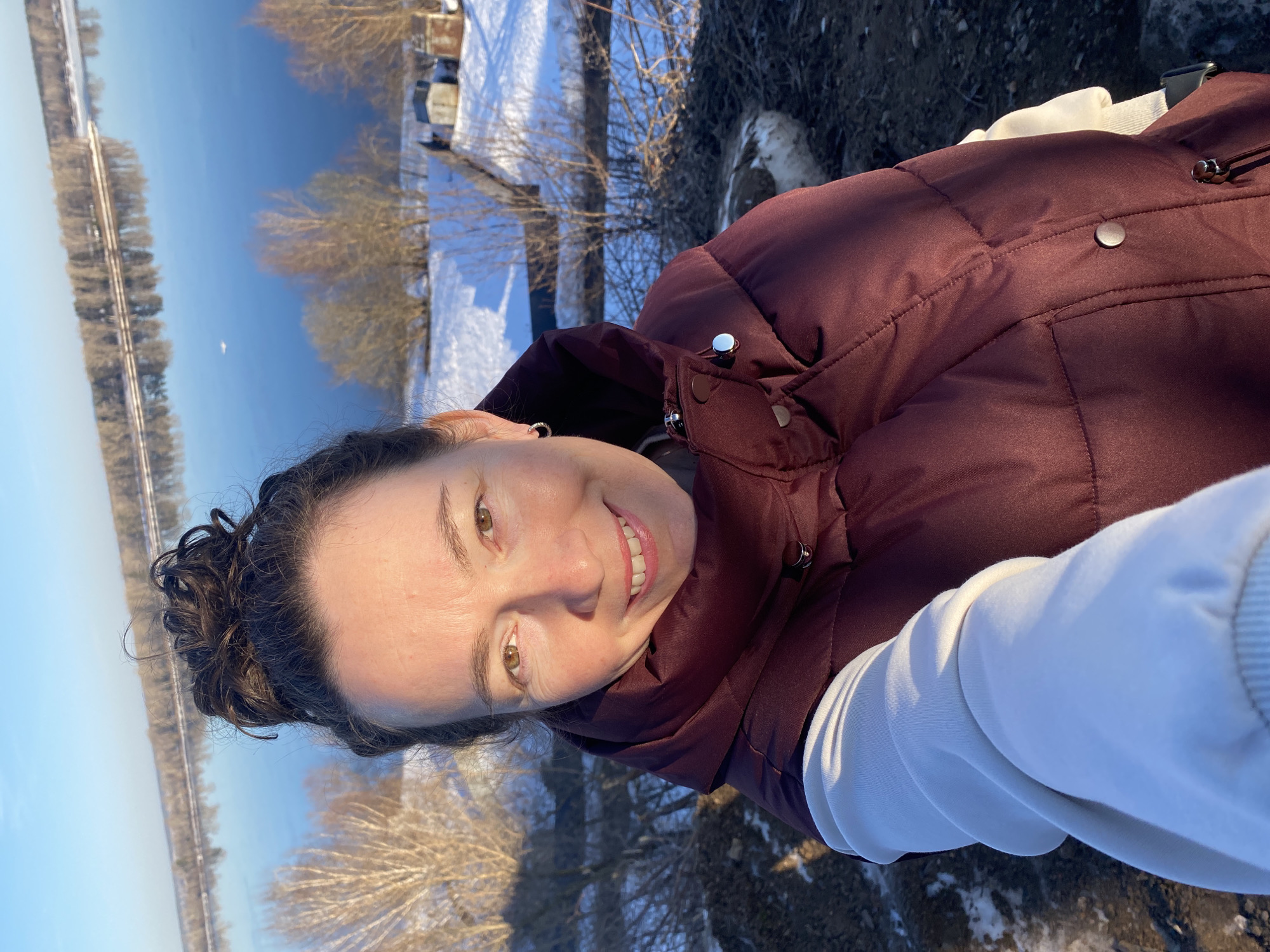A woman in a maroon puffer jacket and white sweatshirt takes a selfie in Ольховка, Russia, with a view of a partially frozen river and snow-covered banks in the background. Bare trees are visible along the riverbank and patches of snow are present on the ground. The sky is clear and blue.