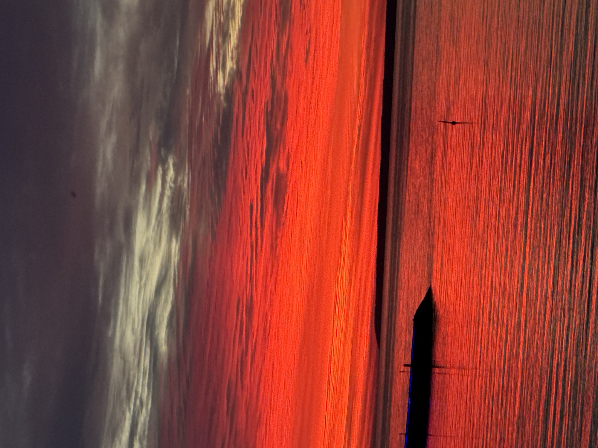 The image depicts a vibrant sunset over the Baltic Sea, as seen from the coast of Tallinn, Estonia. The sky is dominated by intense reds and oranges, with streaks of lighter colors and darker clouds at the top. The sea reflects the fiery hues of the sky, creating a mirror-like surface with visible ripples. In the foreground, a dark, angular structure, possibly a pier or a breakwater, juts out into the water from the left side of the frame. A thin, dark vertical object, likely a buoy or a marker, stands in the water to the right of the pier. The horizon line is defined by a dark band of land in the distance. The overall impression is one of a dramatic and colorful natural spectacle.