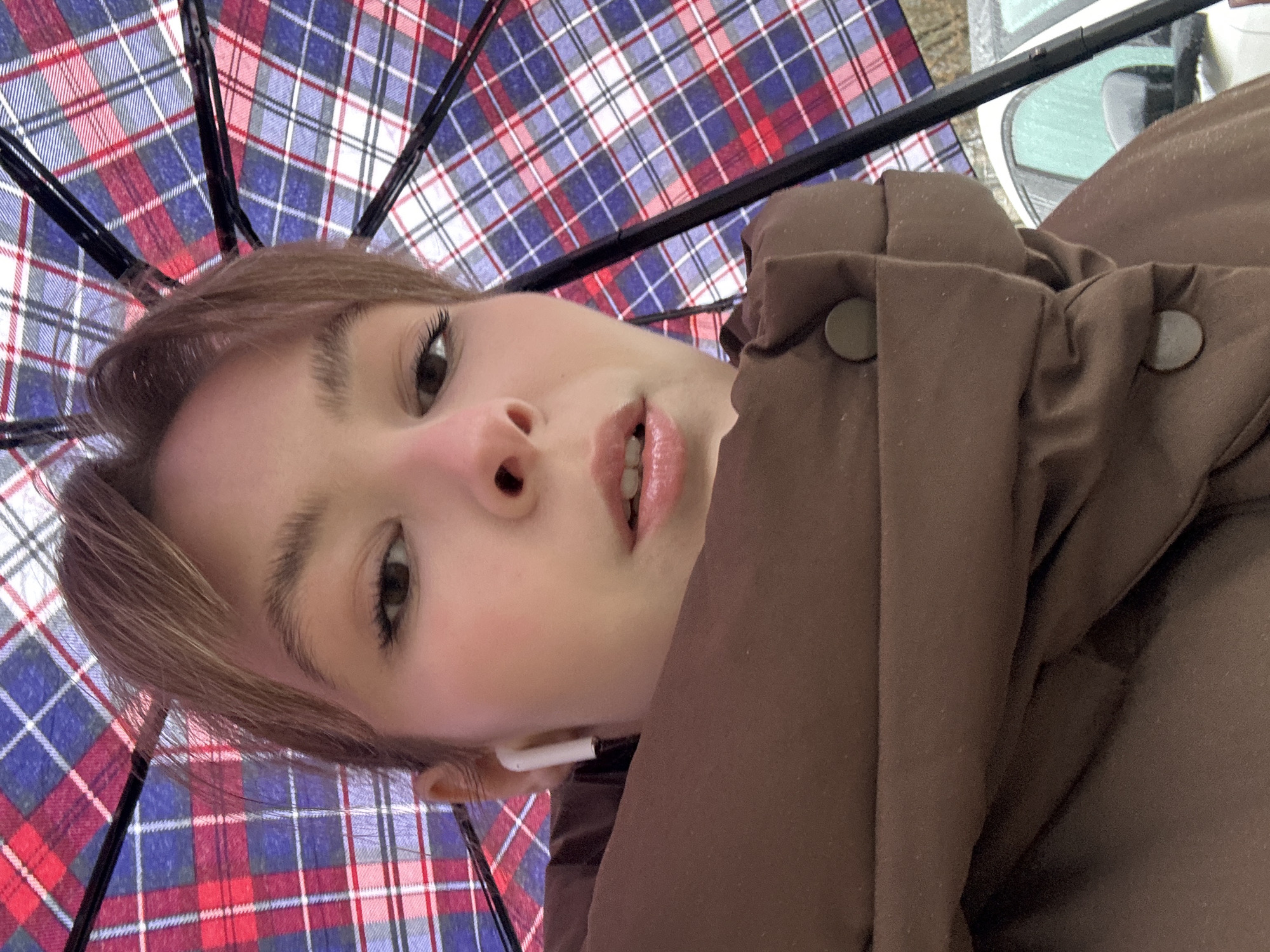 The image is a unique upward-facing selfie of a young woman, captured from a low angle and slightly rotated. The woman is positioned in the lower-left to central part of the frame, looking upwards and slightly to her right. She has light brown hair, dark eyes with visible eyelashes, and glossy, light pink lips that are slightly parted, revealing a glimpse of her upper teeth. A white wireless earbud is visible in her left ear.
She is wearing a brown puffer jacket or coat, characterized by its thick, padded texture and visible rounded buttons or snaps near the collar area.
The dominant background element is the interior of an open umbrella, featuring a distinctive tartan or plaid pattern in red, blue, and white, with dark support ribs radiating outwards. Above the umbrella, a small, blurred section suggests an outdoor environment, possibly a reflection from a window or a distant street scene, though no specific details are discernible. The lighting suggests it is daytime. The overall mood is casual. No visible text is present in the image. The scene is set in what could be Chaikovsky, Russia.