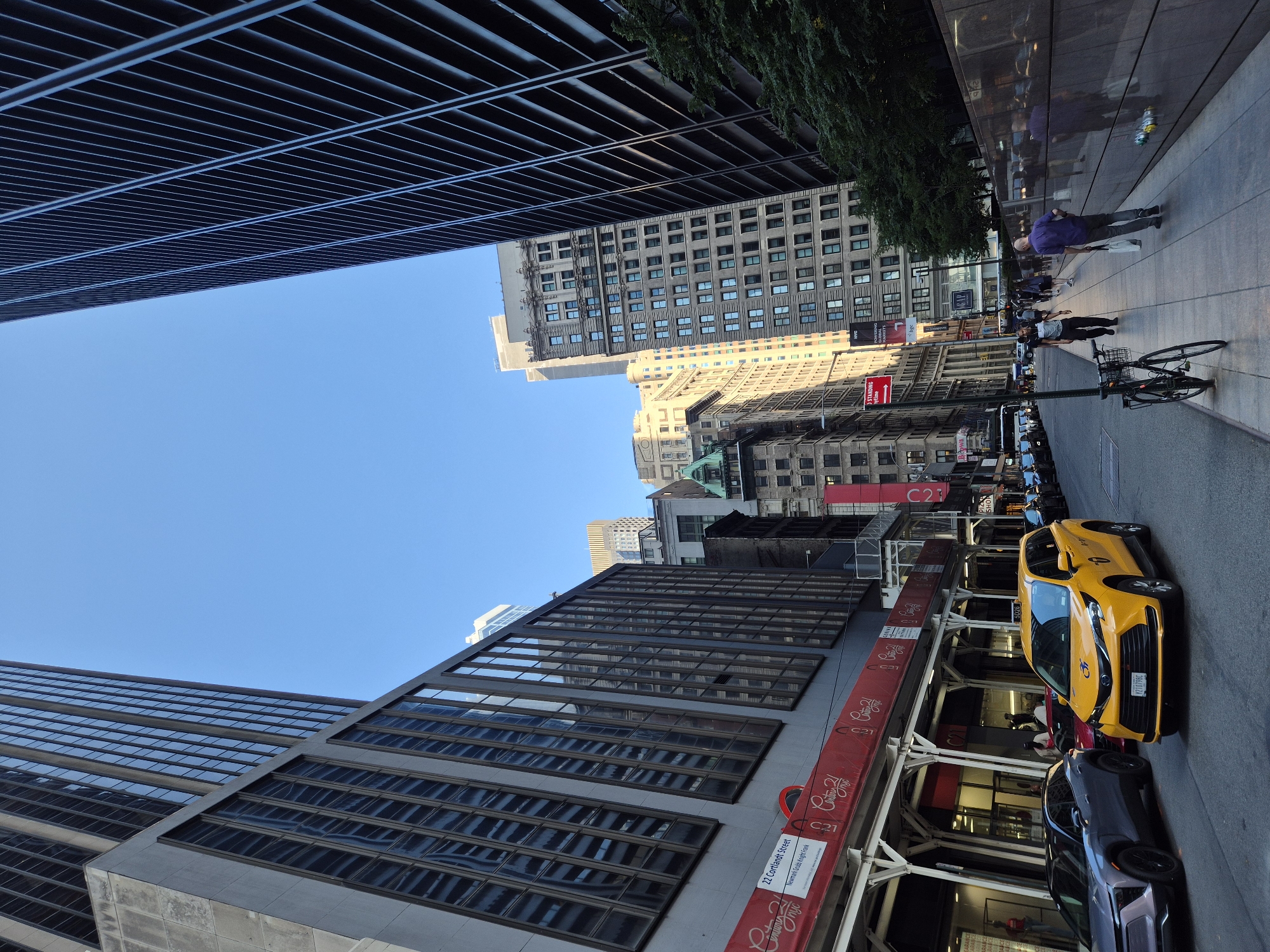 A street scene in New York City, United States of America, depicts a sunlit urban environment with towering buildings. The perspective is low, looking up at skyscrapers that flank a street. On the left, a gray building with dark, reflective windows dominates the foreground. A red banner with white text "Century 21" is visible along its base, partially obscured by scaffolding. A yellow taxi with a license plate "Y210798C" is stopped in the street, next to a dark gray SUV. Pedestrians walk along a sidewalk on the right, where a dark, reflective wall lines the street. One man in a purple shirt carries a white bag, while another walks ahead in a dark shirt and pants. A bicycle is parked on the sidewalk. Further down the street, more buildings of varying architectural styles and heights are visible, with a clear blue sky above. The scene suggests a bustling commercial district on a bright day.