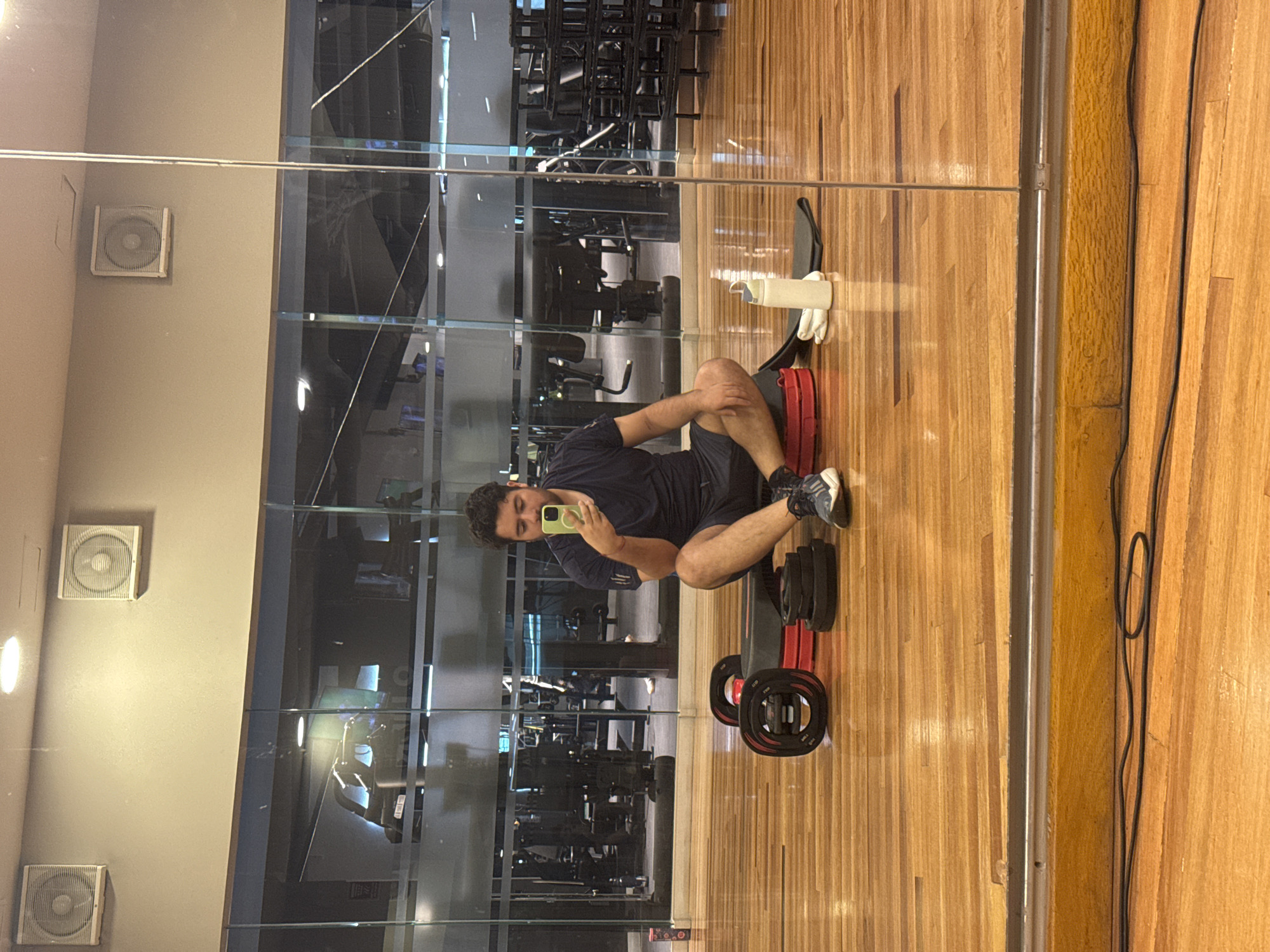 An image captures a young adult male taking a selfie while seated on the wooden floor of a gym in Puebla, Mexico. The man, likely in his 20s or 30s, has short dark hair and is wearing a dark blue t-shirt, dark shorts, and athletic shoes. He holds a light-colored smartphone, directing his gaze towards it to capture his reflection in a large mirrored wall.
The setting is an indoor fitness center with a polished wooden floor. The prominent mirrored wall reflects an array of gym equipment, including weight machines and racks, suggesting a busy exercise area beyond the immediate frame. To the left of the mirrored wall is a plain, light-colored wall featuring three square ventilation grilles, indicating an active air circulation system.
Near the man, on the wooden floor, are several pieces of gym equipment: black barbell plates with red accents, a red and black exercise step or bench, a white water bottle, and a pair of white workout gloves. Black electrical wires or cables run vertically along the right edge of the floor, close to a metal strip that separates the wooden floor from another section or wall.
The activity clearly shows the man taking a break, engaging with his phone in a casual pose. The lighting is artificial and bright, typical of an indoor gym environment. No discernible text is visible anywhere in the image.