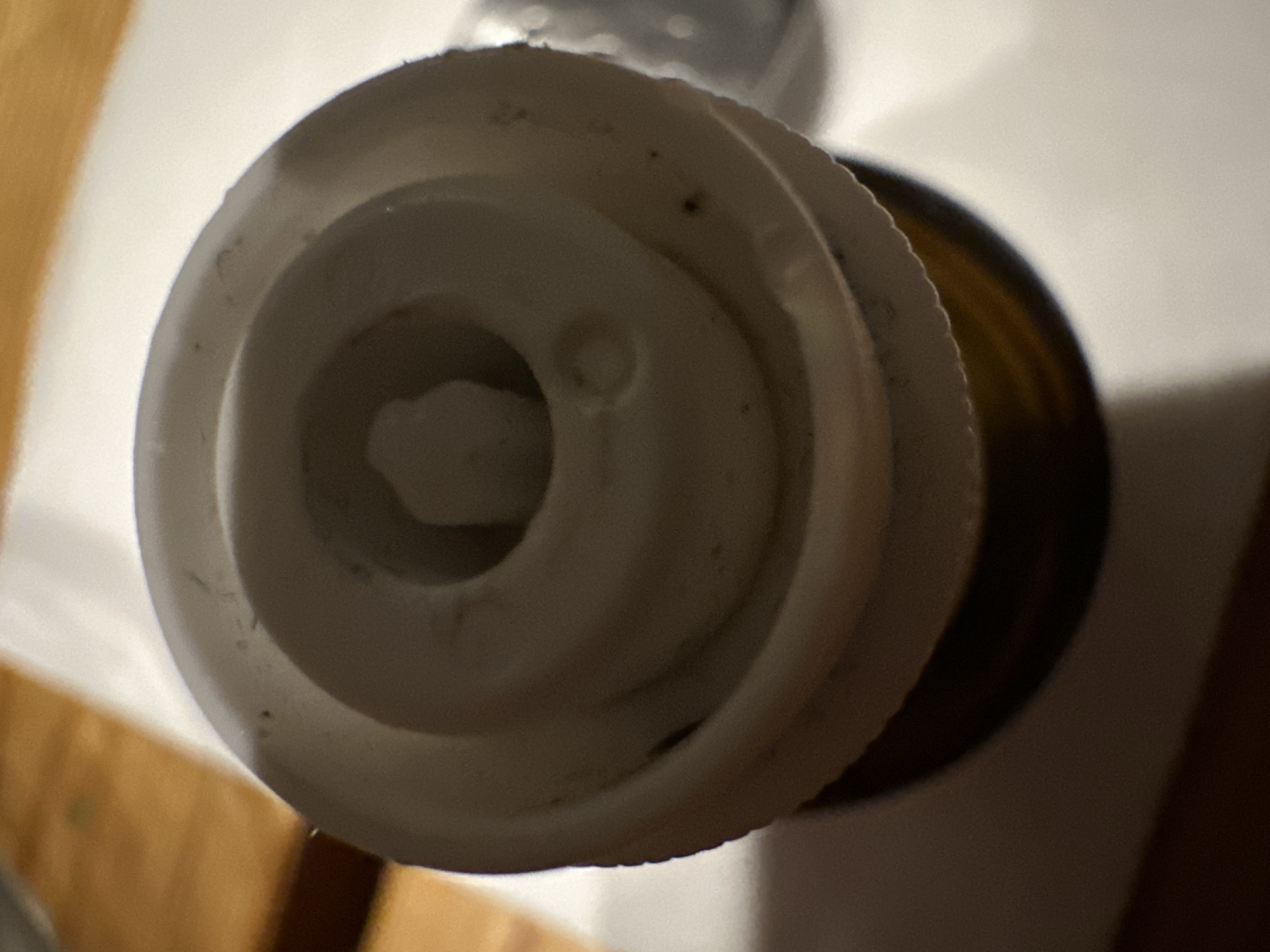 The image shows a close-up, top-down view of the cap of a brown glass bottle, likely containing a liquid or powder. The cap is made of white plastic with a concentric ring design. A small, raised projection is visible in the center of the cap. The background is blurred, but appears to be a wooden surface on the left and a white surface on the right. There are a few dark specks, possibly dirt or debris, on the cap and the surrounding surfaces. The bottle and its cap are the sole focus of the image, with no other objects or individuals present. The setting is indoors, with no indication of the specific location within Востец Снт, Russia, beyond what can be inferred from the general appearance of the objects.