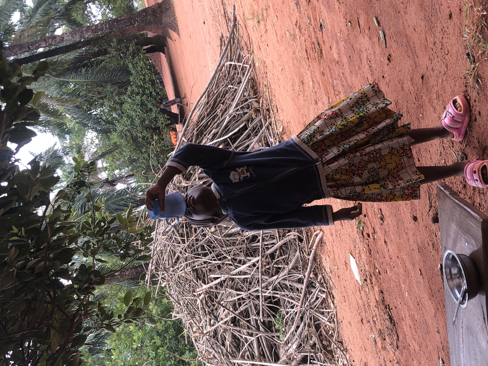 A young girl stands in an outdoor setting in Nteje, Nigeria. She wears a dark blue long-sleeved shirt, a colorful floral skirt, and pink sandals. She holds a blue cup or container near her head. Behind her is a large pile of thin branches or sticks. The ground is reddish-brown dirt. In the background are trees and greenery, including what appears to be palm trees. To the right, a small table or platform with a metallic bowl and utensil on it is visible. The lighting indicates it is likely daytime.