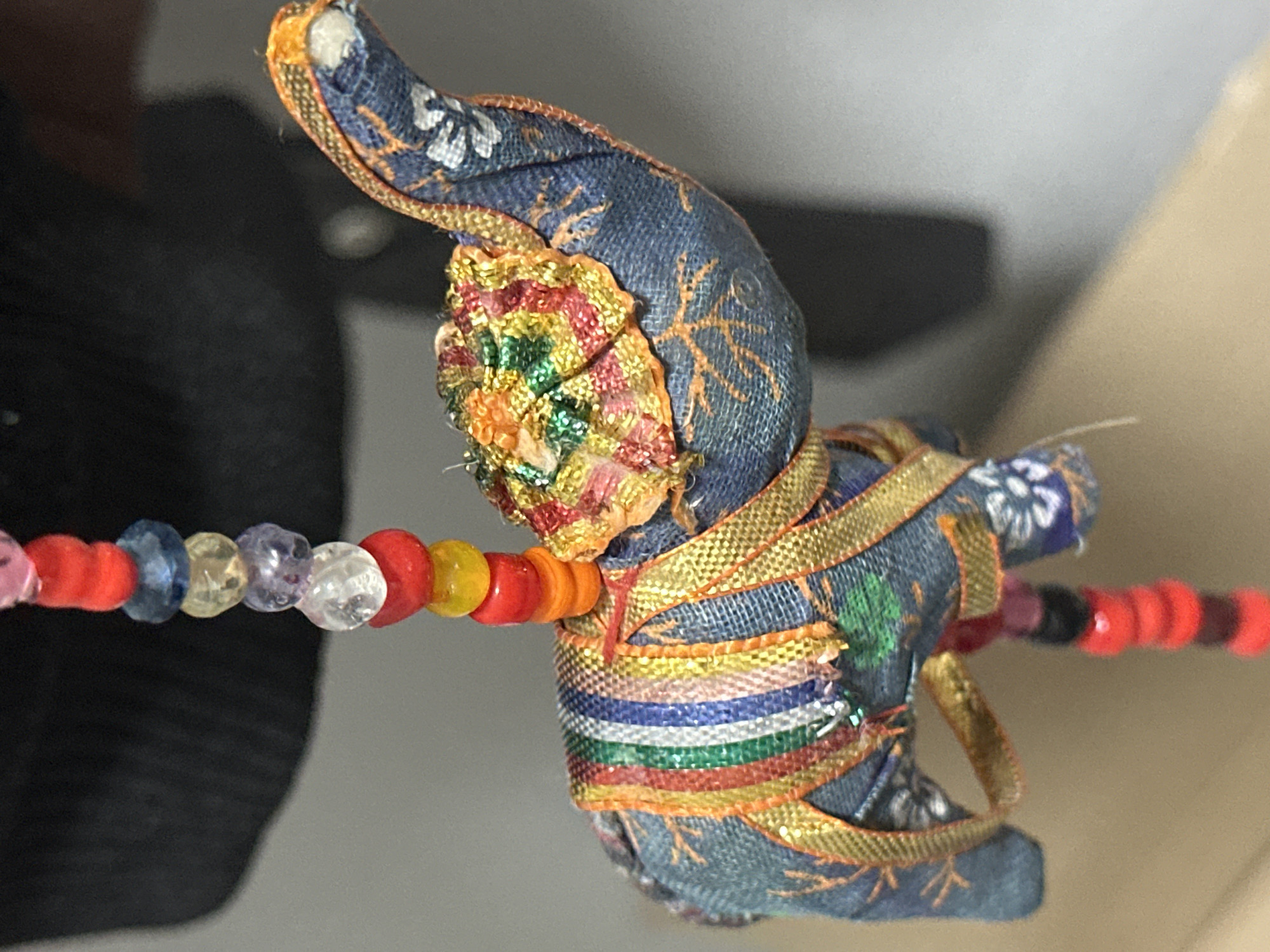 A close-up, indoor shot features a small, fabric-made elephant ornament hanging by a beaded string. The elephant is adorned with colorful ribbons and intricate beadwork on its back. The background is softly blurred, suggesting an indoor setting with ambient light. The image does not contain any elements that would allow for identification of a specific location in Nevinnomyssk, Russia.