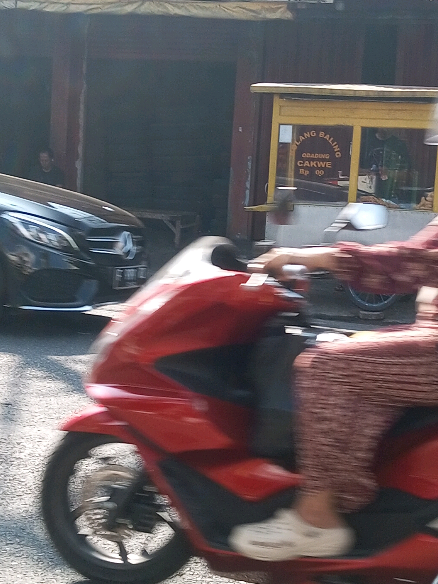 The image shows a street scene in Wonosobo, Indonesia, with a focus on a red scooter in the foreground, moving from left to right. A person is riding the scooter, with their legs and lower torso visible. They are wearing a patterned skirt and white slip-on shoes. Behind the scooter, a black car with a Mercedes-Benz logo is parked. Further in the background, there is a food stall with a yellow awning, displaying text that appears to be in Indonesian, including "LANG BALING", "ODADING", and "CAKWE". A person is partially visible inside the stall. The environment appears to be an outdoor urban setting with a paved street and buildings. The image is slightly blurred due to motion.