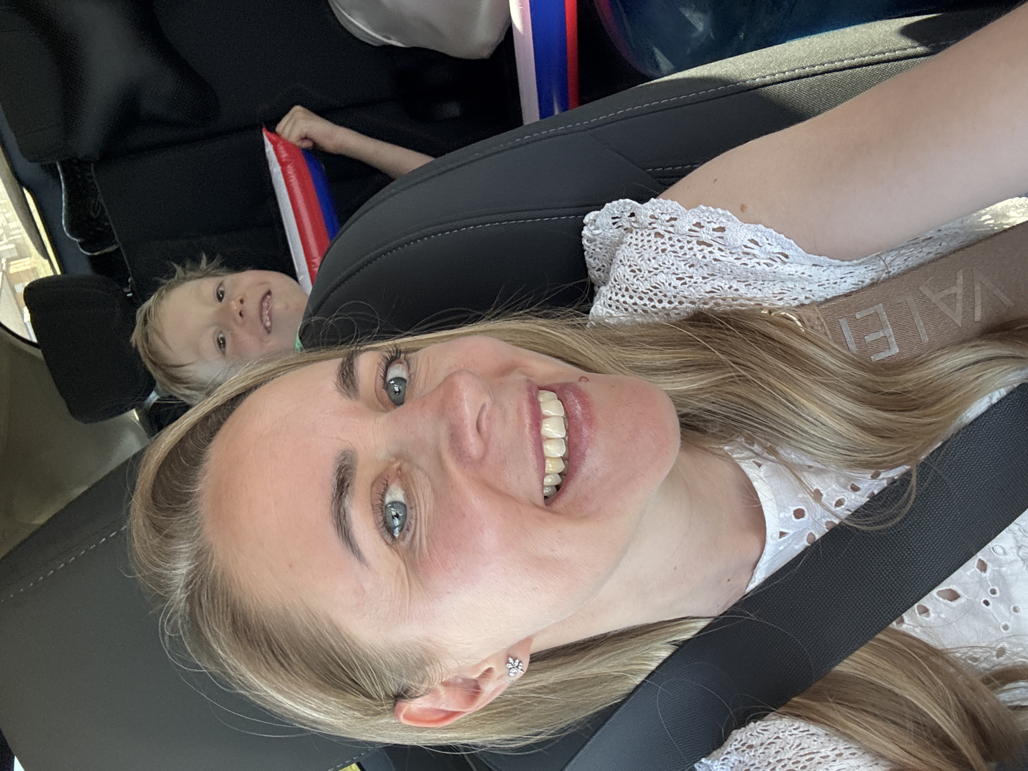 A medium-shot selfie shows a woman smiling at the camera while wearing a seatbelt in what appears to be a car. The woman has light blonde hair, blue eyes, and is wearing a white lace top. In the background, a child's face is visible, and the child is holding an inflatable stick with red, white, and blue colors, resembling the Russian flag. The interior of the car is visible, with black leather seats. The context suggests the location could be Chaikovsky, Russia, given the flag colors.