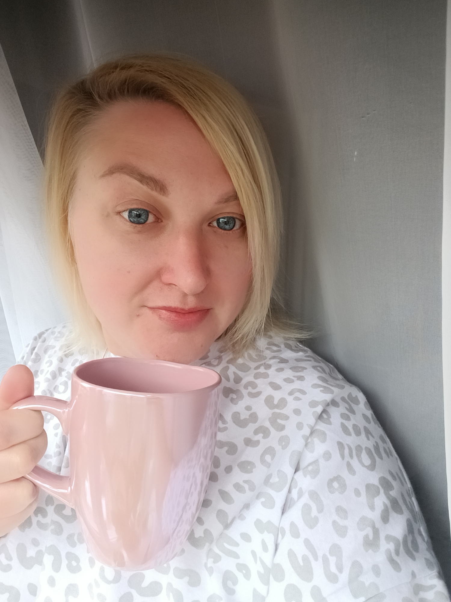A fair-skinned woman with blonde hair and blue eyes is holding a pink mug. She is wearing a white shirt with a light grey animal print. She appears to be standing indoors, possibly near a window with white sheer curtains and a grey wall. There is no verifiable information to determine the location to be Chaikovsky, Russia.