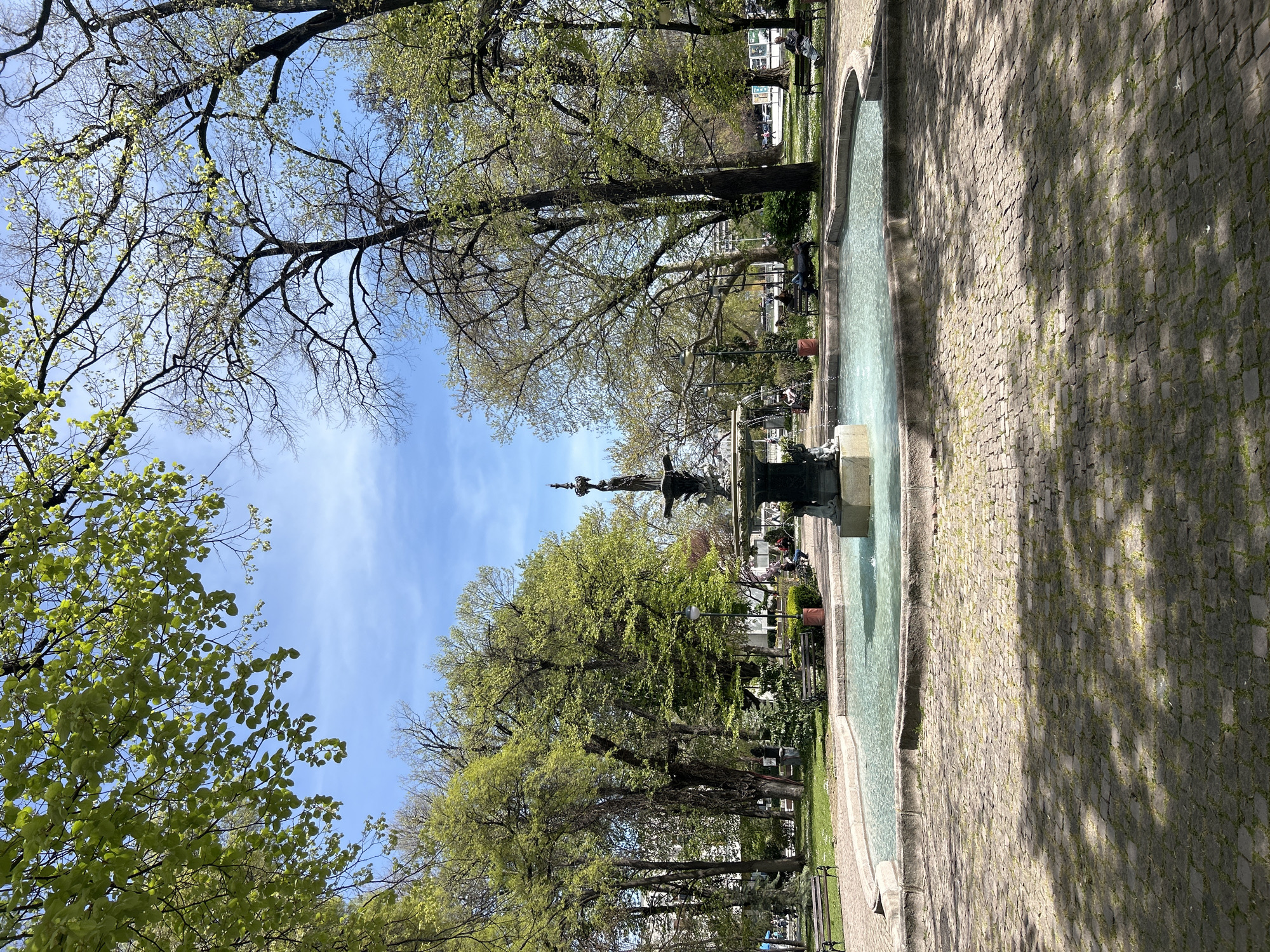 This image captures a serene park scene in Plovdiv, Bulgaria, showcasing the arrival of spring. The photograph is oriented vertically, depicting a long, narrow public space flanked by trees and featuring a prominent water element.
Numerous trees dominate the scene, with many displaying fresh, light green foliage, indicating early or mid-spring. Some upper branches still appear bare against the clear, vibrant blue sky, which has a few subtle white clouds.
A long, rectangular water basin or channel runs along the right side of the central pathway. Within this basin, approximately midway through the frame, a multi-tiered fountain sculpture stands, actively spraying water and contributing to the tranquil atmosphere. The water in the basin is a light blue-green, reflecting the sky.
Adjacent to the water feature on the right is a wide pathway paved with uneven cobblestones. Sunlight filters through the tree canopy, casting distinct shadows and bright patches onto the ground. On the left side, beyond the water feature, are grassy areas interspersed with more trees and several dark-colored park benches. A few individuals are visible, some sitting on these benches, enjoying the pleasant weather. A lamp post can be seen near the fountain.
The overall impression is one of a peaceful, sunny daytime setting, likely morning or early afternoon, with people relaxing amidst the burgeoning greenery of a well-maintained urban park. There is no visible text in the image.