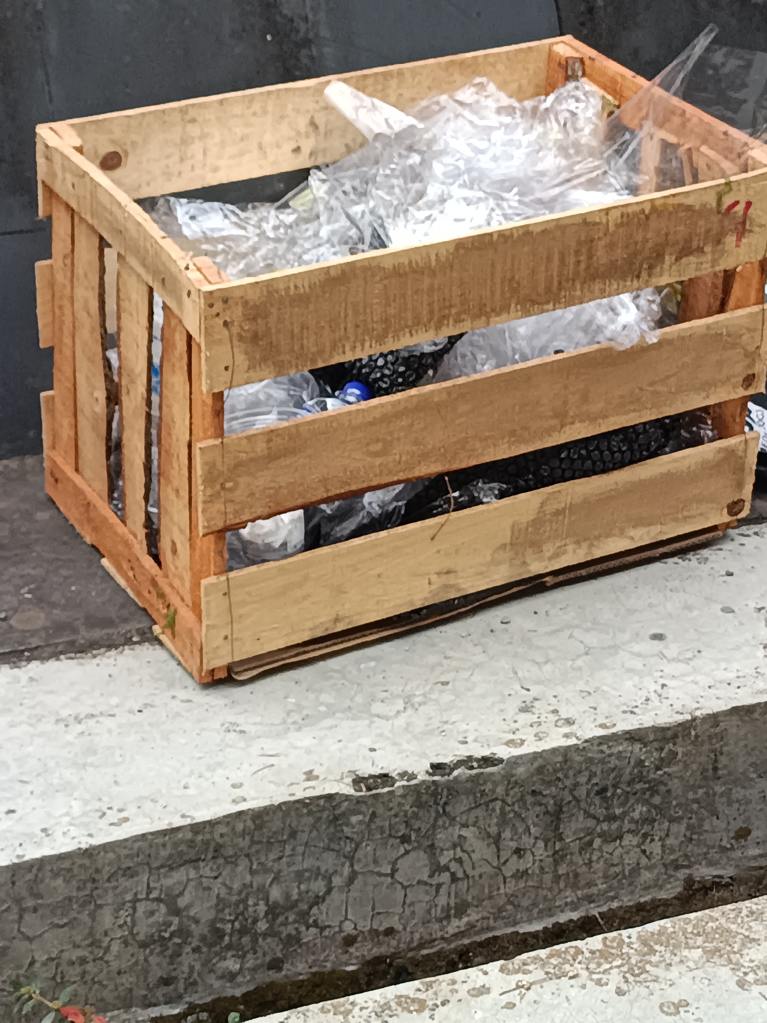 A wooden crate filled with plastic bags and dark, granular material sits on a cracked concrete surface. The crate is constructed of rough-hewn wooden planks, with some of the planks appearing weathered and slightly damaged. Inside the crate, clear plastic bags are visible, some containing crumpled plastic items and others filled with the dark, granular substance. The crate is positioned outdoors, with a dark, textured wall in the background and a rough concrete pathway in the foreground. The lighting suggests it is daytime. The scene appears to be a part of an urban or semi-urban environment in Hargobinangun, Indonesia, possibly an area where waste is collected or stored. There are no people or animals visible in the frame.
