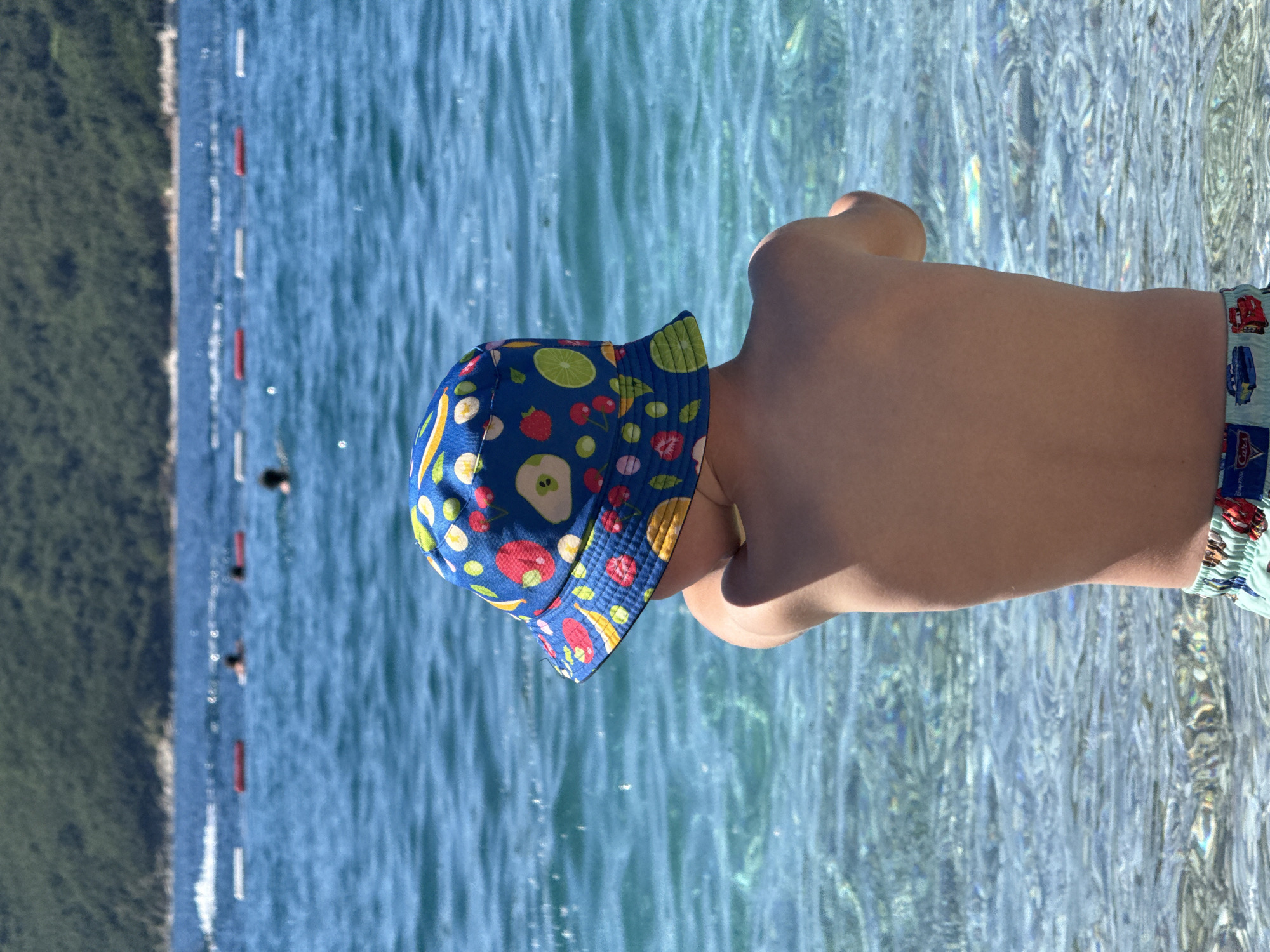 A young child, viewed from the back, stands in the shallow, clear turquoise water off the coast of Budva, Montenegro. The child wears a blue fruit-patterned sun hat and "Cars" themed swim trunks. The water shimmers in the sunlight. In the background, a coastline with vegetation is visible, along with a swimming area delineated by red and white buoys. Several people are swimming further out in the water. The image is taken from a slightly elevated perspective.