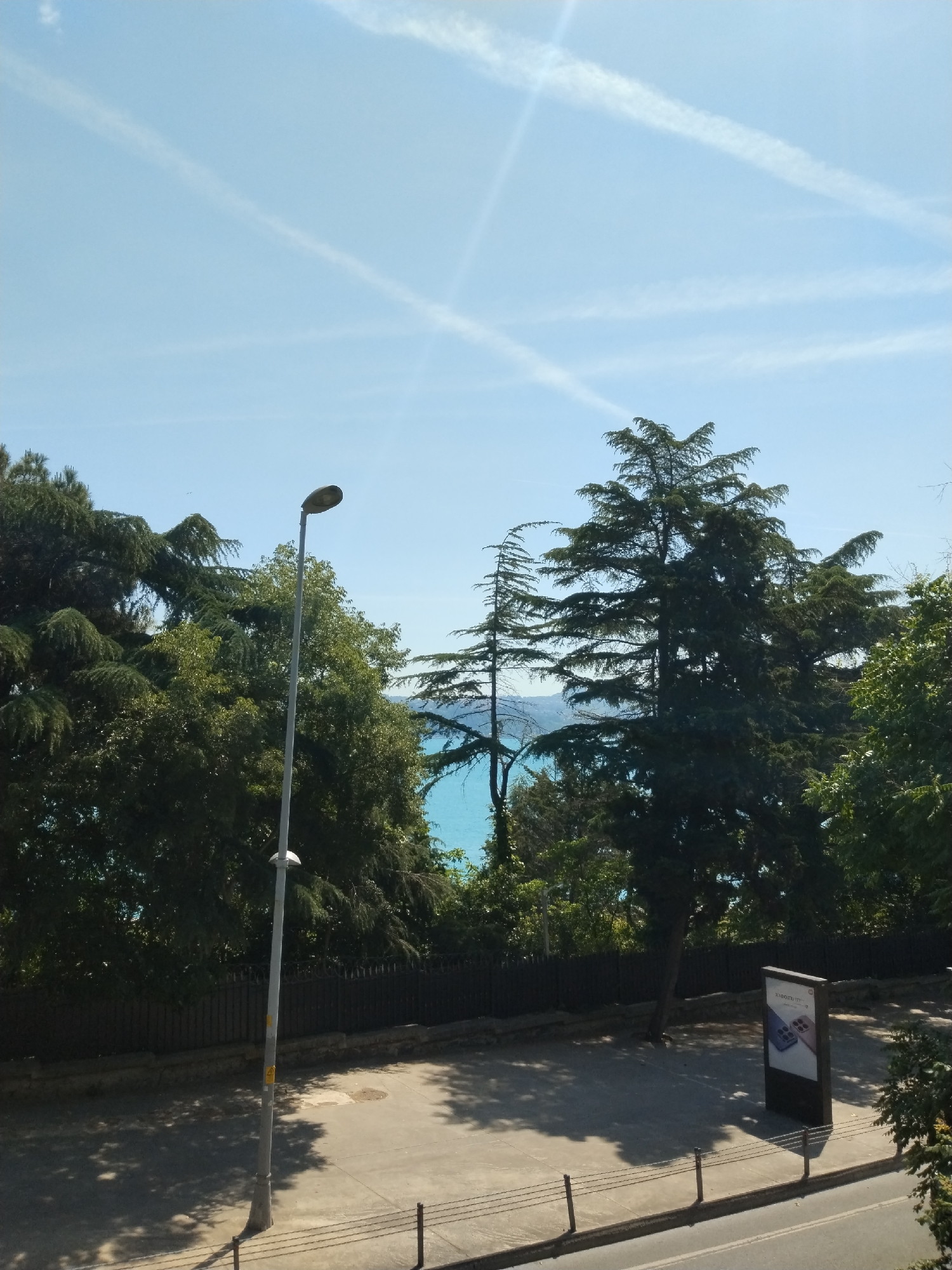 A vibrant outdoor scene in Sarıyer, Turkey, showcases a juxtaposition of natural beauty and urban elements under a bright, clear sky. The upper portion of the image features a pale blue sky streaked with several distinct white contrails, indicating recent air traffic.
The midground is dominated by a lush canopy of dense, dark green trees, predominantly coniferous, with varied textures. Through a visible gap in the foliage towards the center-right, a striking expanse of turquoise-blue water, likely the Bosphorus, is glimpsed. A tall, slender grey lamppost stands centrally, reaching towards the treetops.
Below the treeline, a dark metal fence runs horizontally, separating the wooded area from a paved ground. This grey surface, possibly a sidewalk or a small road, exhibits pronounced shadows cast by the surrounding trees and the lamppost, suggesting a sunny day, likely morning or early afternoon. To the right of the paved area, a modern, dark-colored vertical digital advertisement display is prominent. The text "Xiaomi 13 Pro" is clearly legible at the top of the display, which also features images of two mobile phones. In the immediate foreground, a low metal railing with multiple strands lines the edge of the paved path. The scene is devoid of people or animals, conveying a sense of serene natural and urban harmony.
