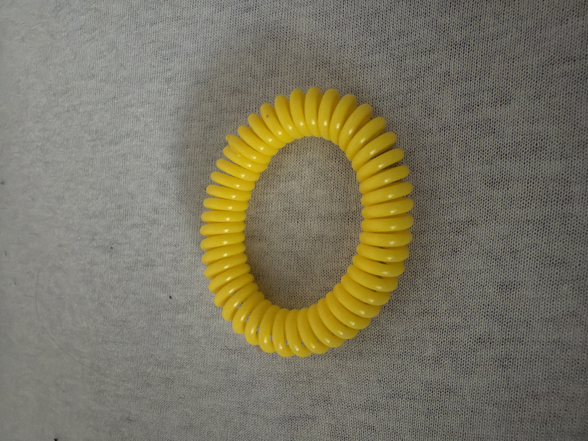 A yellow, coiled plastic bracelet or hair tie is centered on a gray textured surface. The surface appears to be a cloth or fabric. The bracelet is the primary focus. No people or location information are discernible from the image.