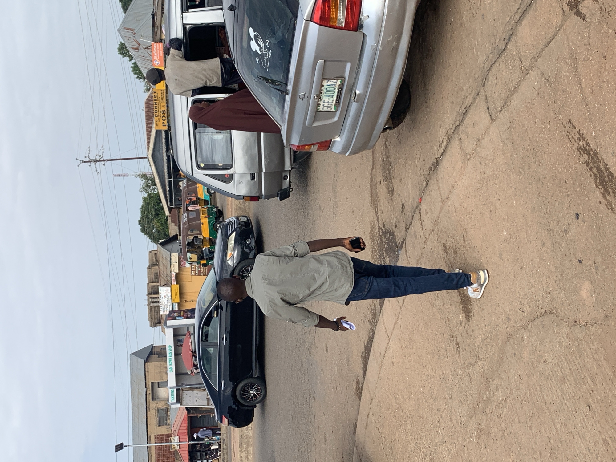 The image depicts a street scene in Gura Top, Nigeria, with a clear view of the "DADI IKOWA BUS STOP" sign. The focus is on a young man walking away from the camera on a paved road. He is wearing a light gray, short-sleeved button-down shirt, blue jeans, and white and beige sneakers. In his right hand, he holds a black smartphone, and in his left, a white folded paper.
Several vehicles are present. A black sedan is parked on the left side of the road. To the right, a silver minivan with its side door open is visible, with two individuals partially visible inside. A person wearing a maroon hijab is standing near the open door of the minivan. Behind the minivan, a silver sedan with a Nigerian license plate that reads "GGE 400AZ" is parked, displaying a Mickey Mouse sticker on its rear window. Further in the background, to the left, are low-rise buildings with signage indicating businesses. To the right, a yellow and green auto-rickshaw is parked. The sky is overcast, and power lines are strung across the scene. The ground is a mix of cracked pavement and dirt.
