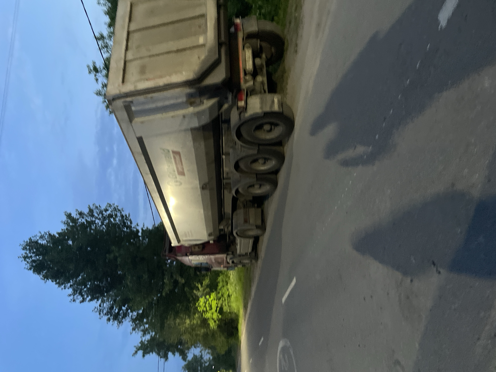A semi-trailer dump truck is parked on the shoulder of a road in Пруды, Russia. The truck has a large, silver-colored trailer and a red cab. The trailer has the name "Goldhofer" printed on its side. The truck is positioned on the right side of the road, facing away from the viewer. The road has white lane markings. In the background, there are trees and lush green vegetation. The sky is a deep blue with some clouds, suggesting it is either early morning or late evening. Two distinct shadows of people are cast on the road in the foreground, indicating their presence or the presence of something casting shadows. The scene appears to be in a rural or semi-rural area on the outskirts of the town.
